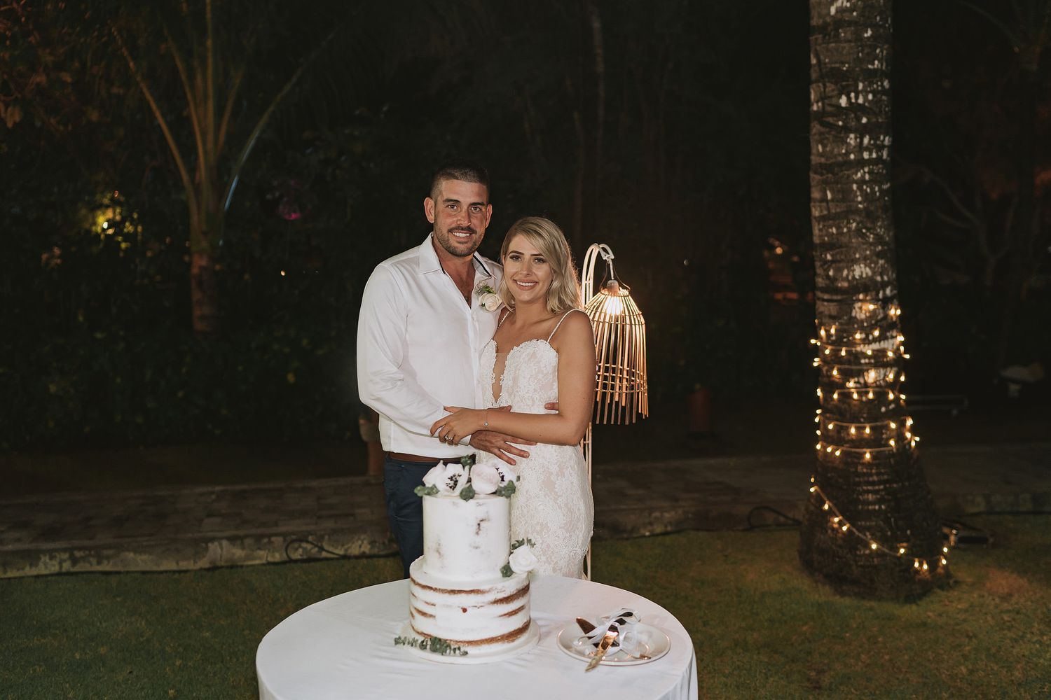Newlywed couple stands behind their two-tiered wedding cake at outdoor evening reception with twinkling lights in background.