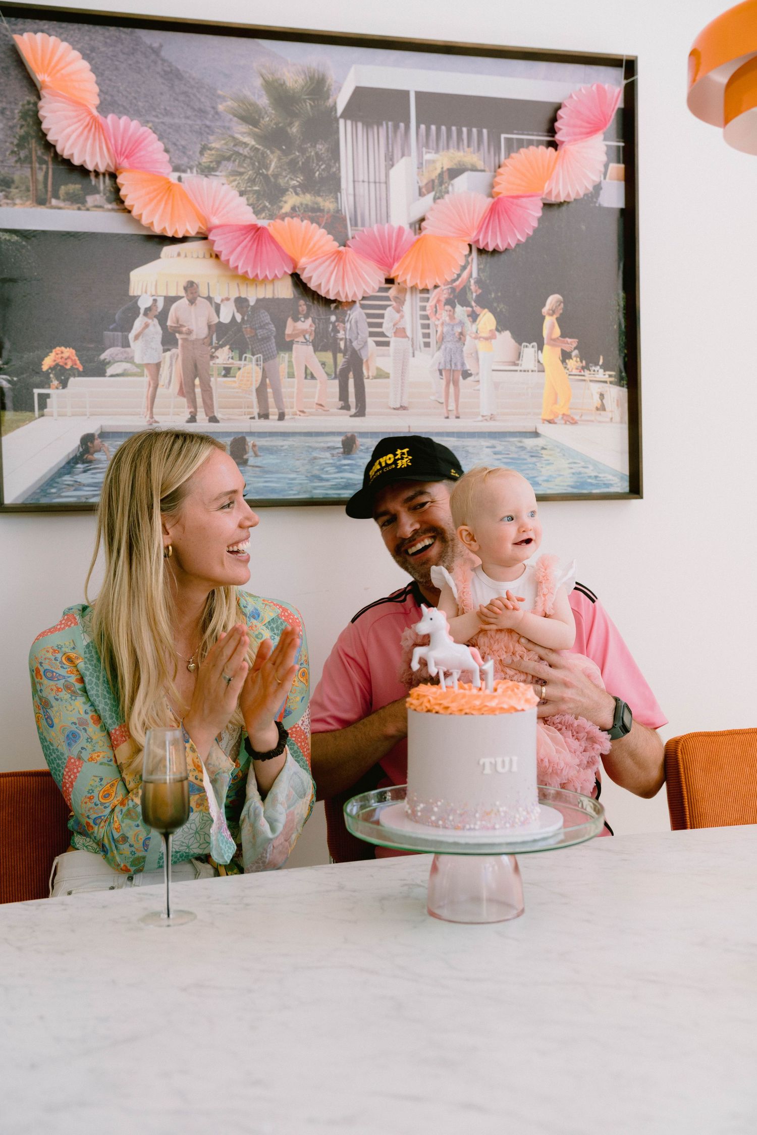 A celebratory moment around a pink birthday cake with coral-colored paper decorations hanging overhead.