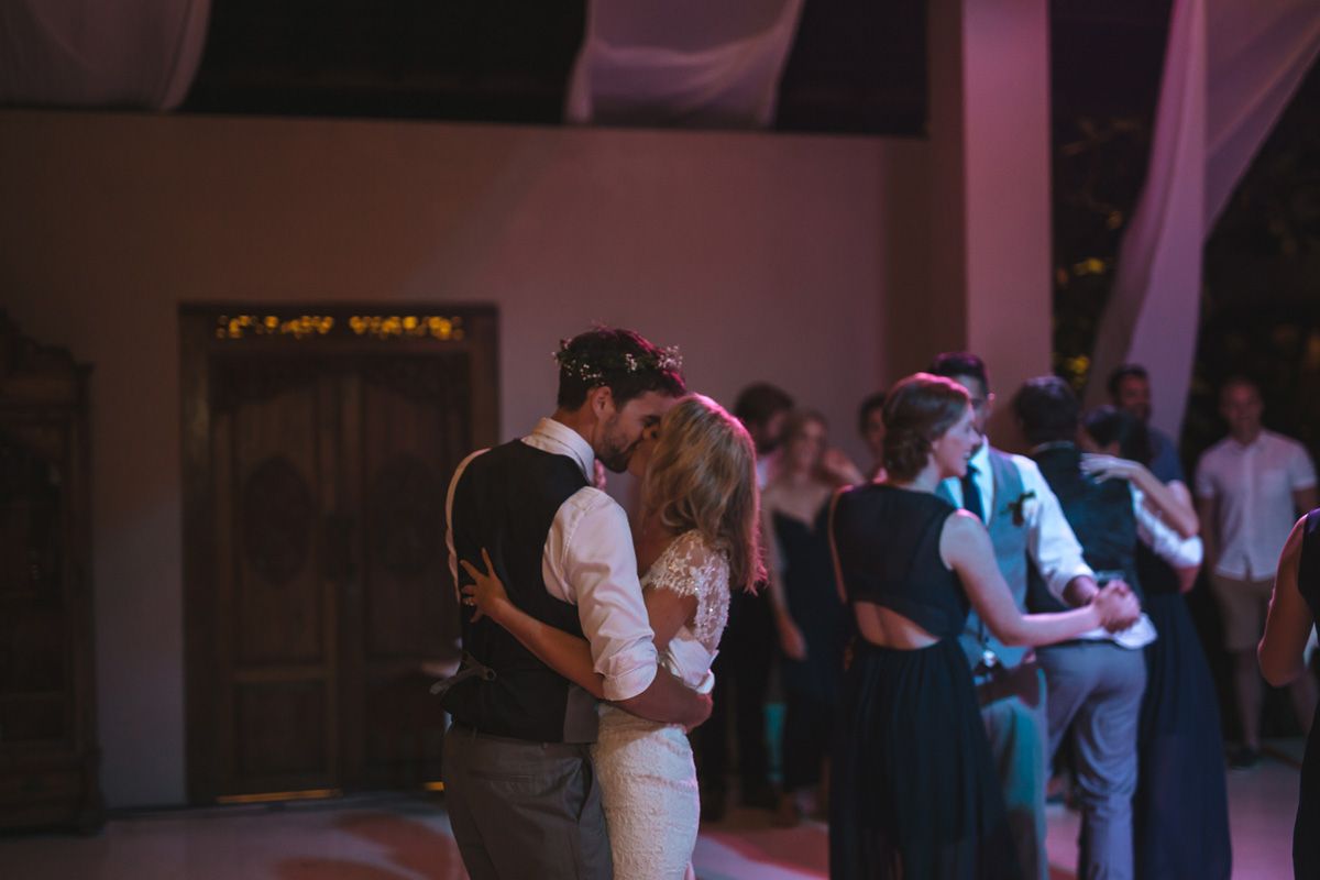Couple sharing a romantic slow dance at a wedding reception with guests in the background under purple lighting.