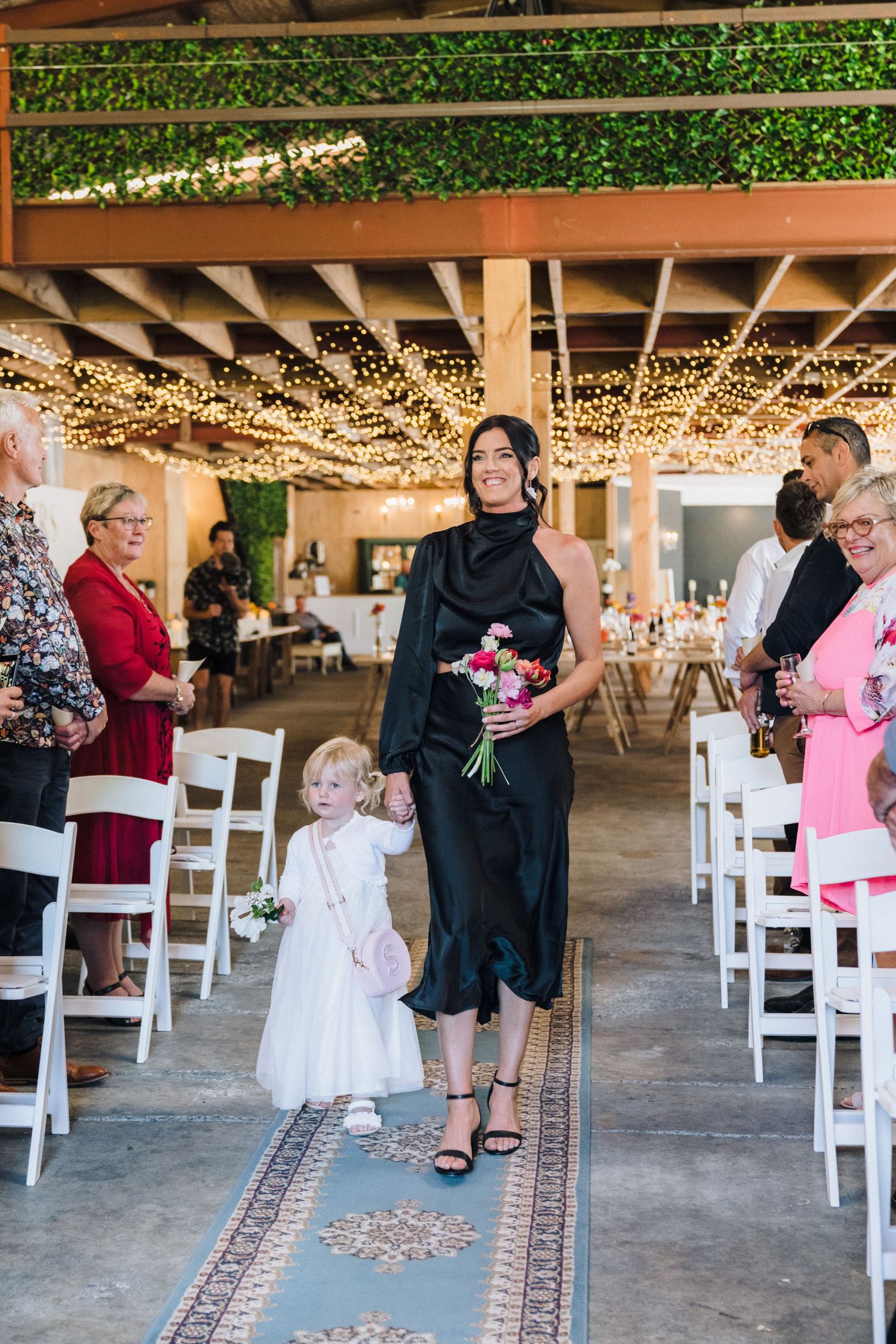 Indoor wedding ceremony setup with string lights, greenery and white chairs creating an aisle.