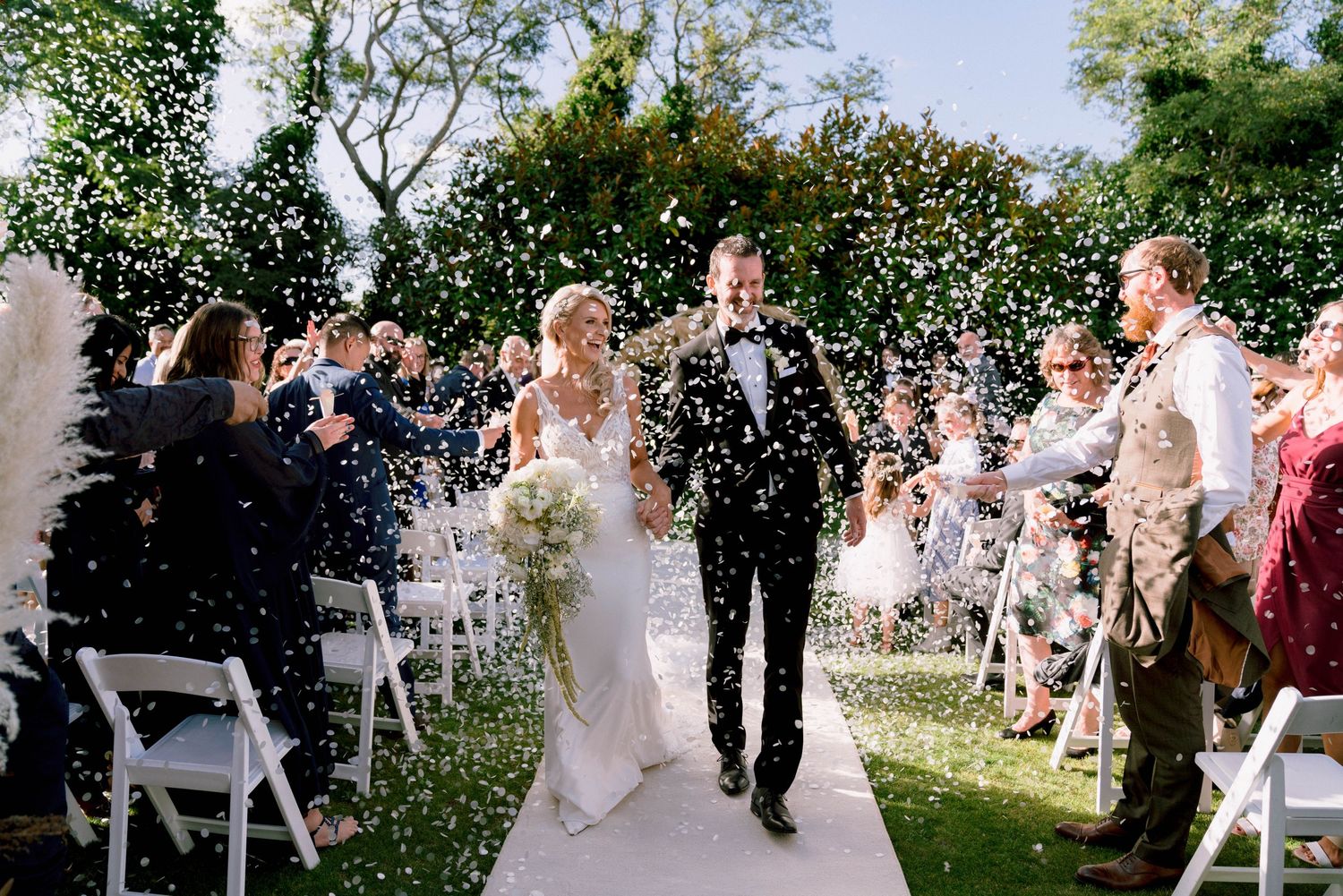 Wedding guests throw white confetti over newlyweds as they walk down the outdoor ceremony aisle on a sunny day.