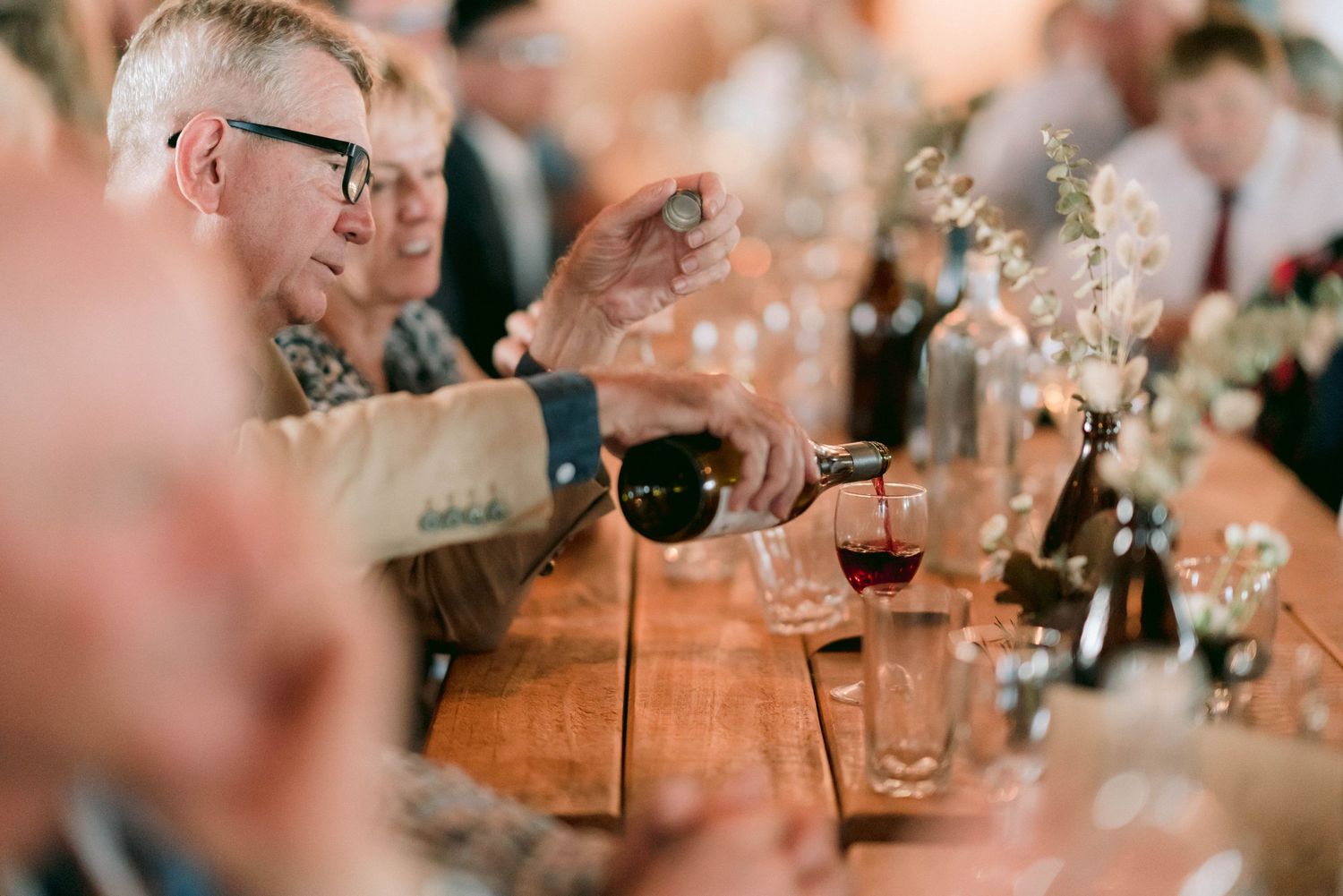 Wedding guests applaud during reception dinner at wooden table with wine glasses.