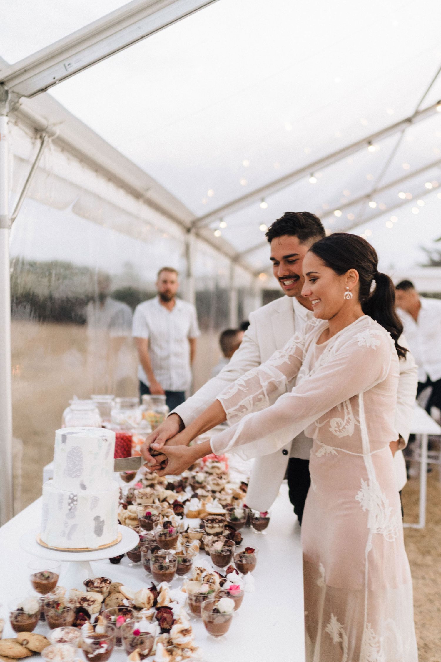 Couple cuts their wedding cake together under white tent with dessert display.