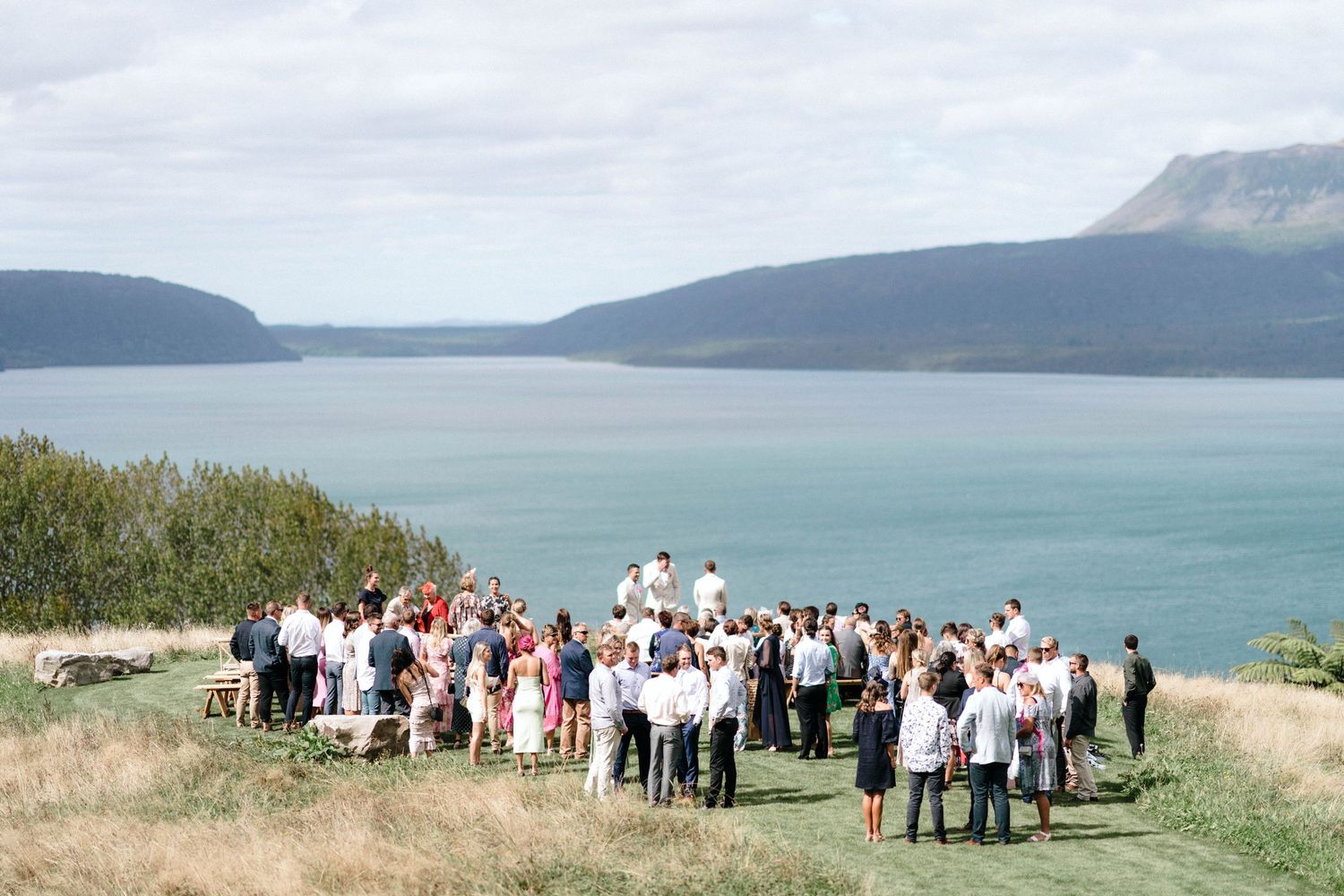 Wedding ceremony guests gathered on a scenic coastal clifftop with mountains and water in the background.