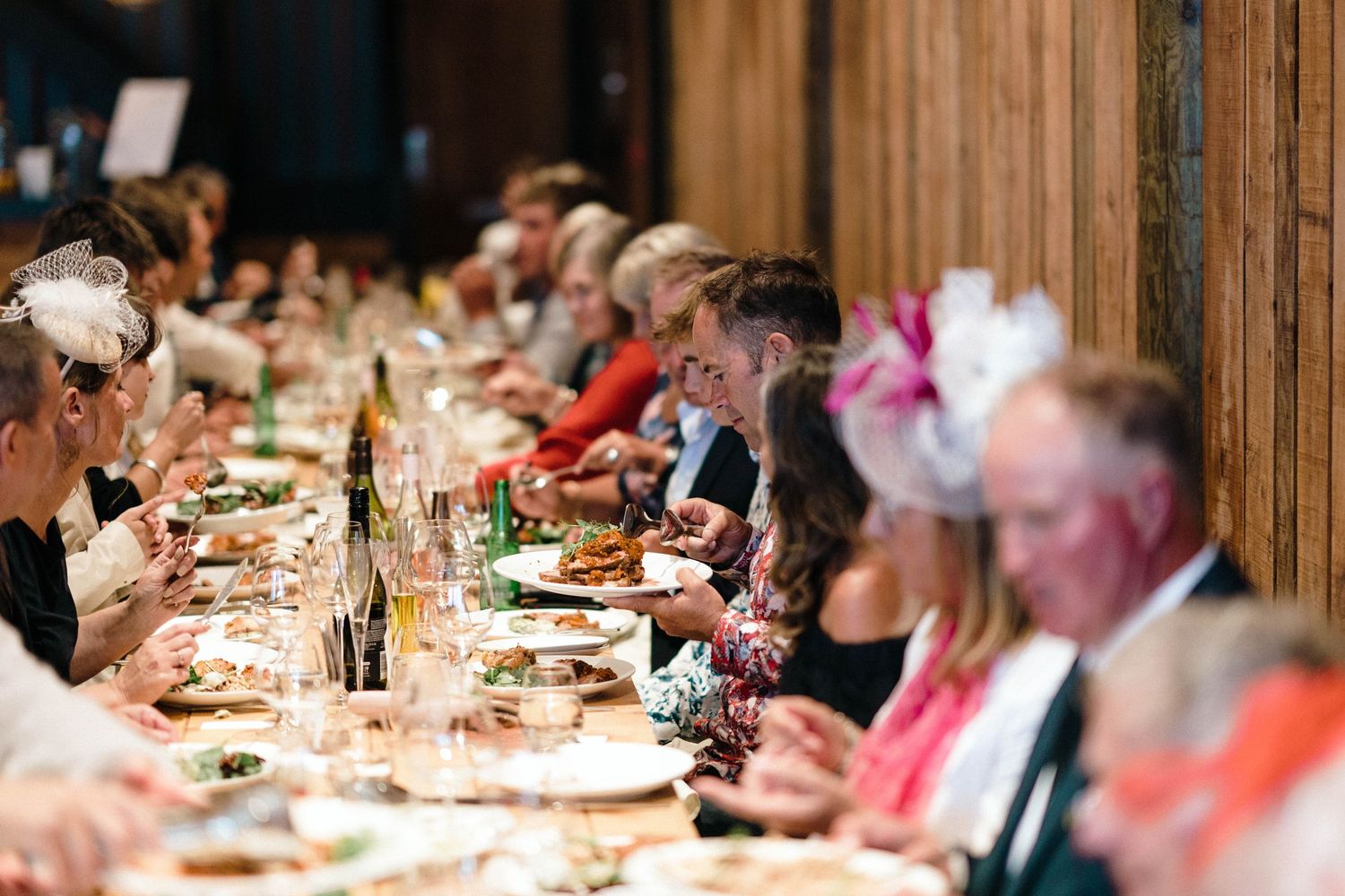 Wedding guests dine at long banquet tables elegantly decorated with candles and floral centerpieces under wooden beams.