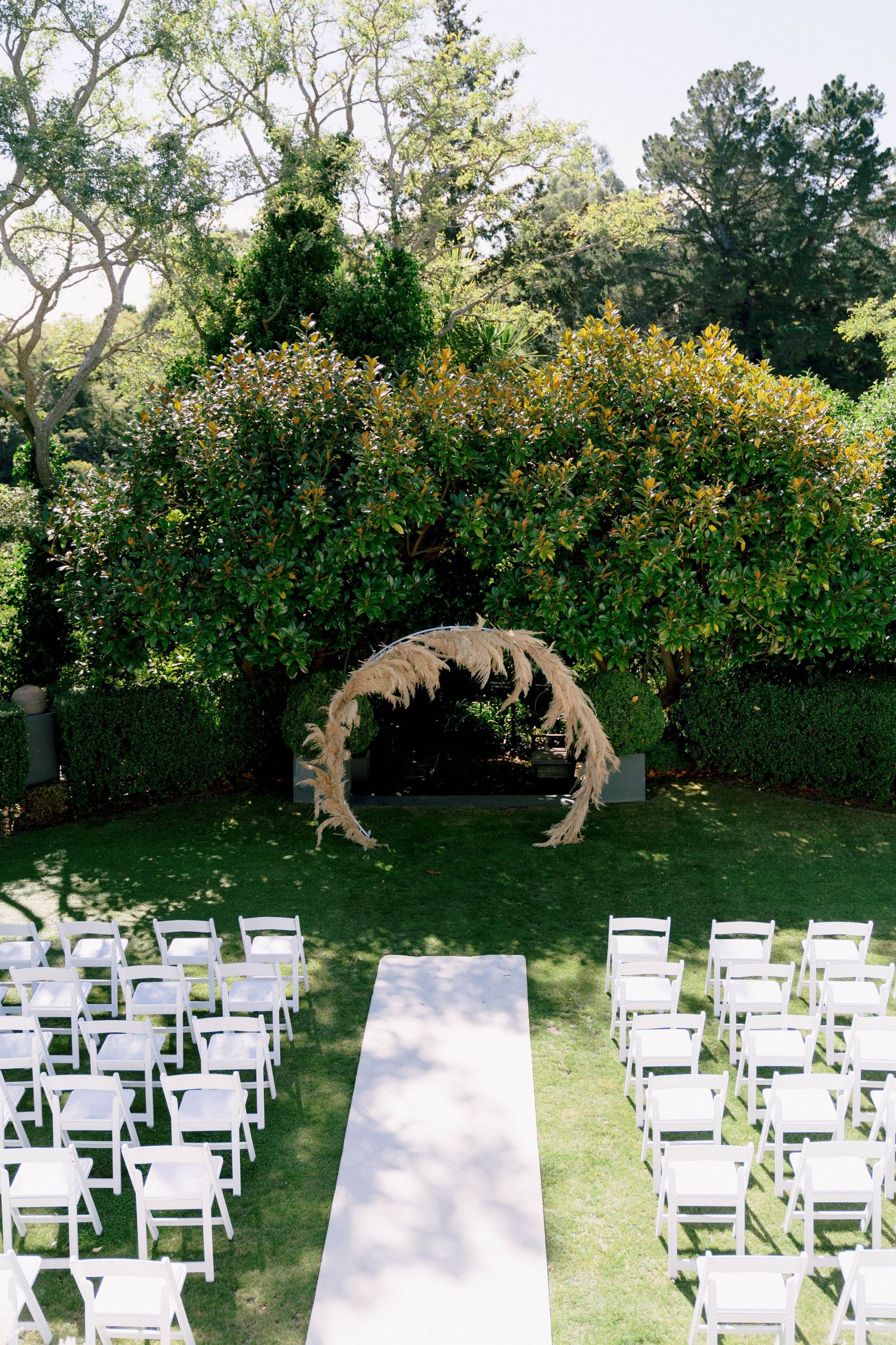 Outdoor wedding ceremony setup with white chairs facing a floral arch and white aisle runner on a manicured lawn.