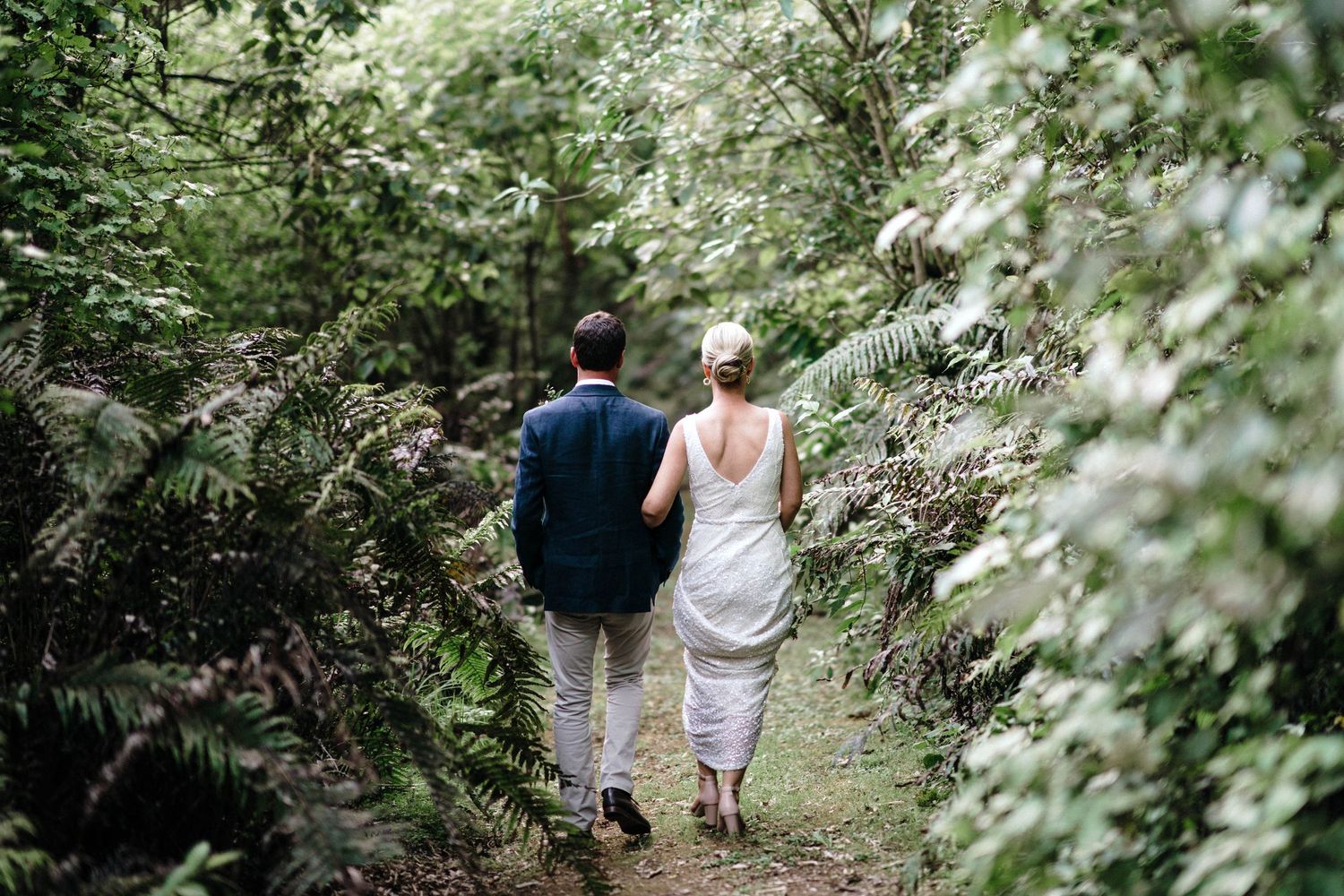 A wedding couple walks down a nature trail surrounded by lush greenery and flowering bushes.