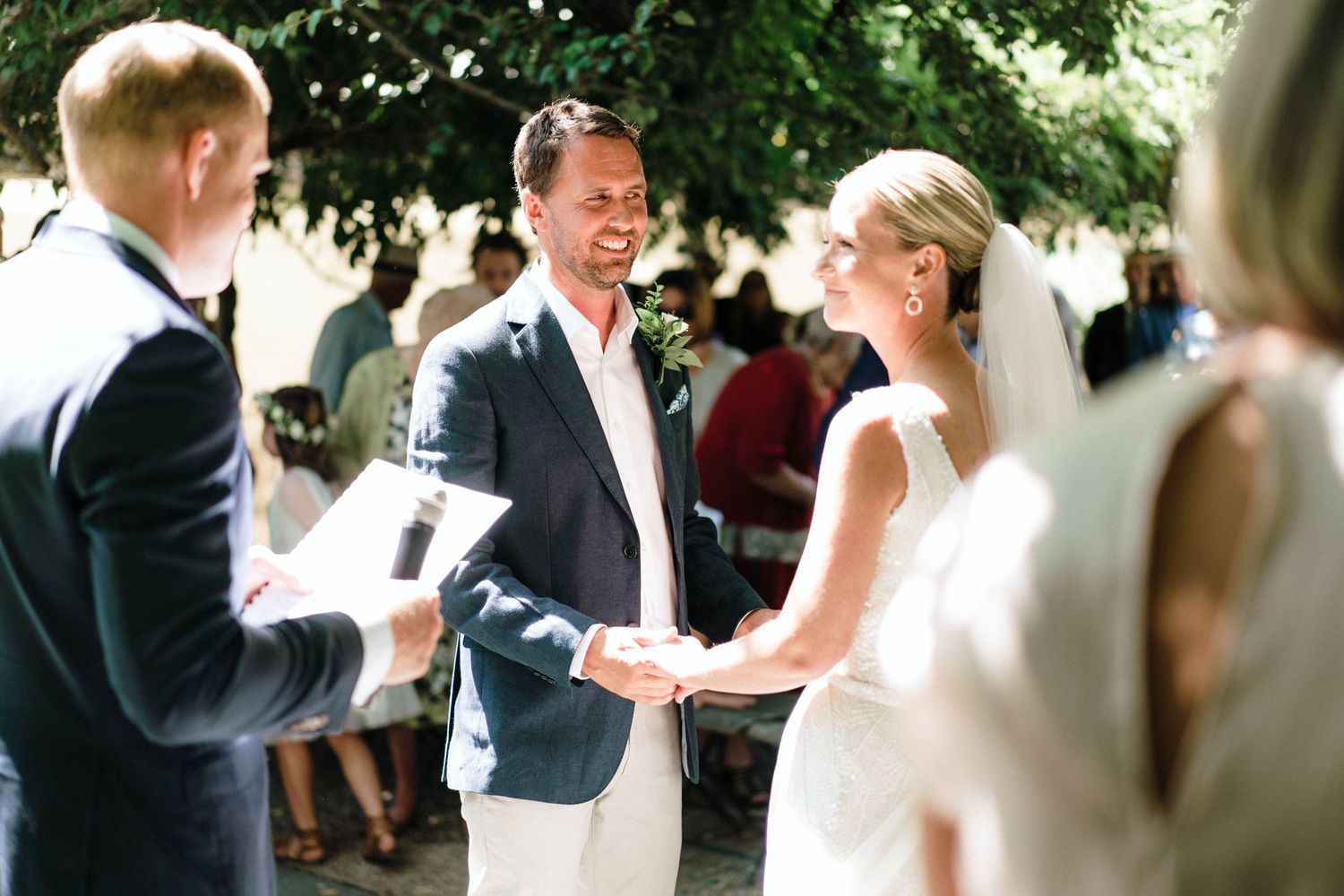 An outdoor wedding ceremony takes place in a garden setting with guests gathered under green trees.