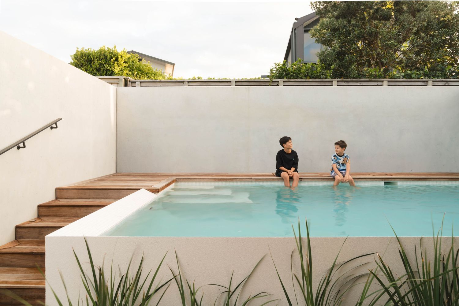 A modern backyard swimming pool with wooden deck stairs and two people sitting on the edge enjoying a sunny day.