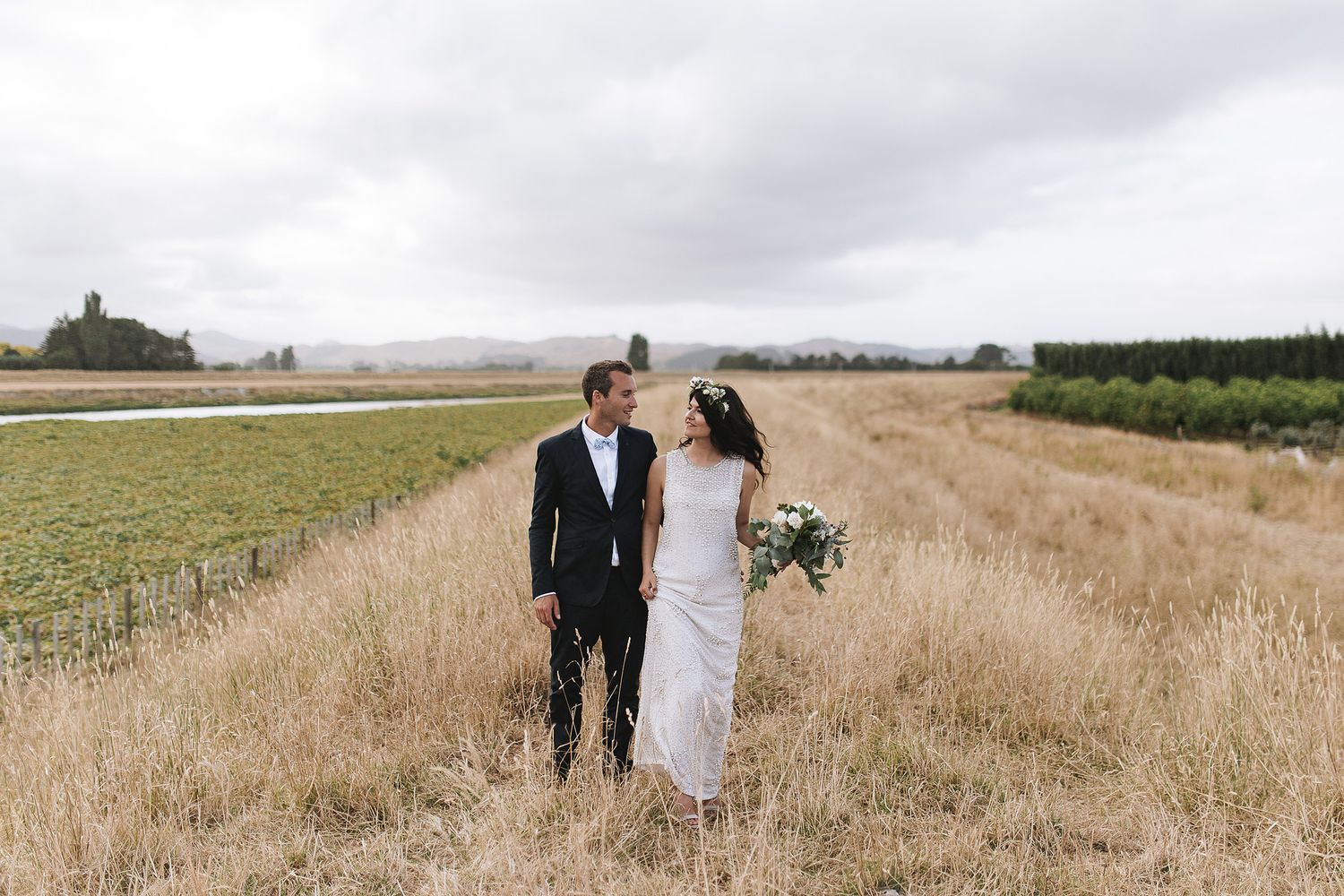 A couple walks through a golden wheat field on a sunny day with rolling hills in the landscape.