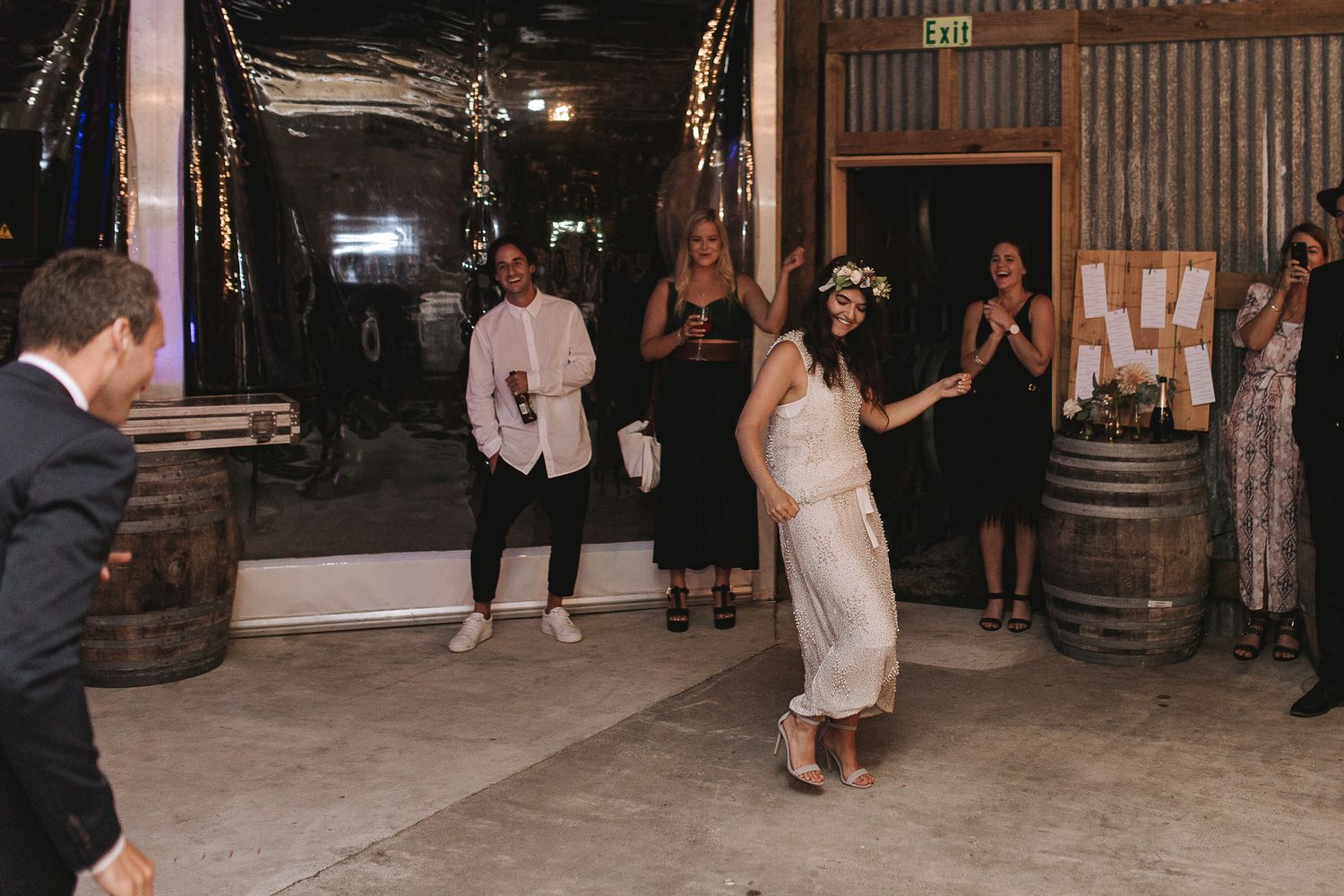 People gather and dance in a rustic barn venue with wooden barrels and corrugated metal walls.
