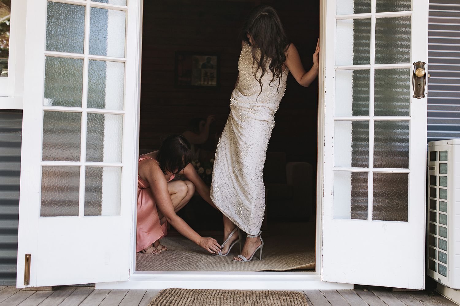 Two people in white dresses playfully pose in a doorway of white French doors opening to a wooden deck.