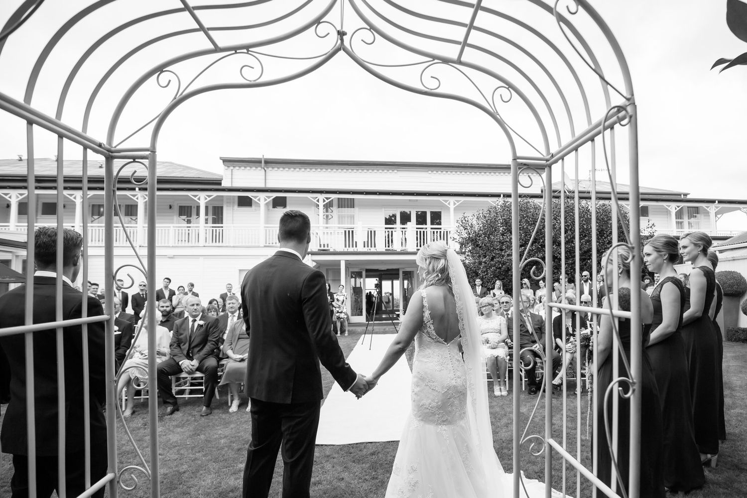 A formal wedding ceremony takes places outdoors with guests seated in white chairs on a sunny day.