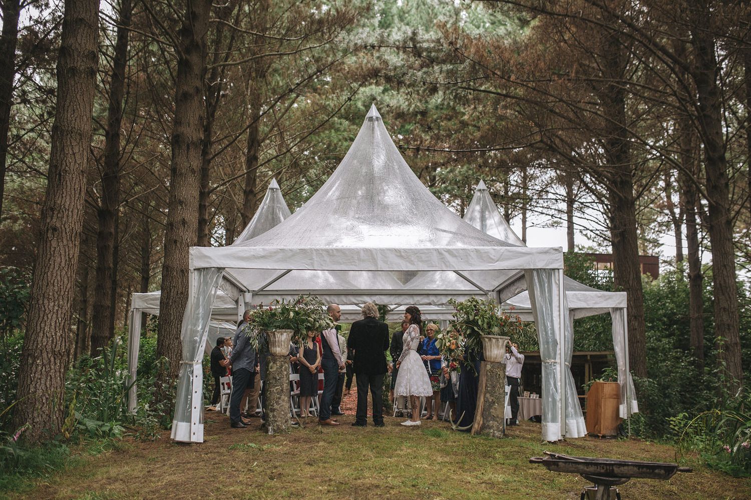 An outdoor wedding ceremony under a white tent pavilion in a forest setting during the day.