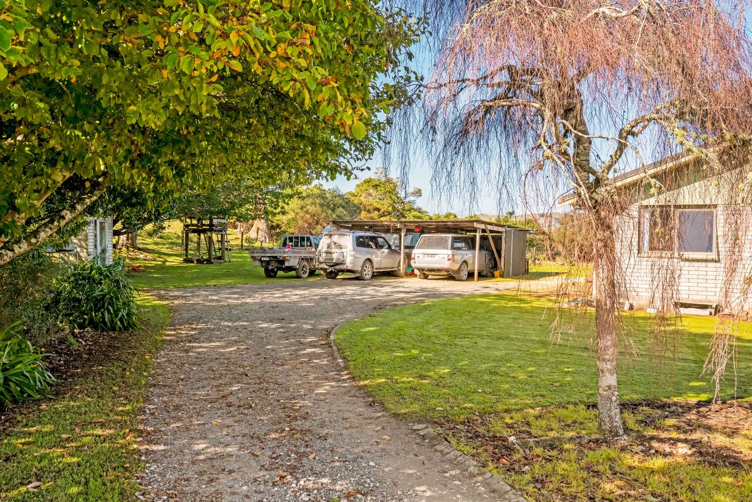 A curved gravel driveway leads to a rural property with parked vehicles and a weeping willow tree.