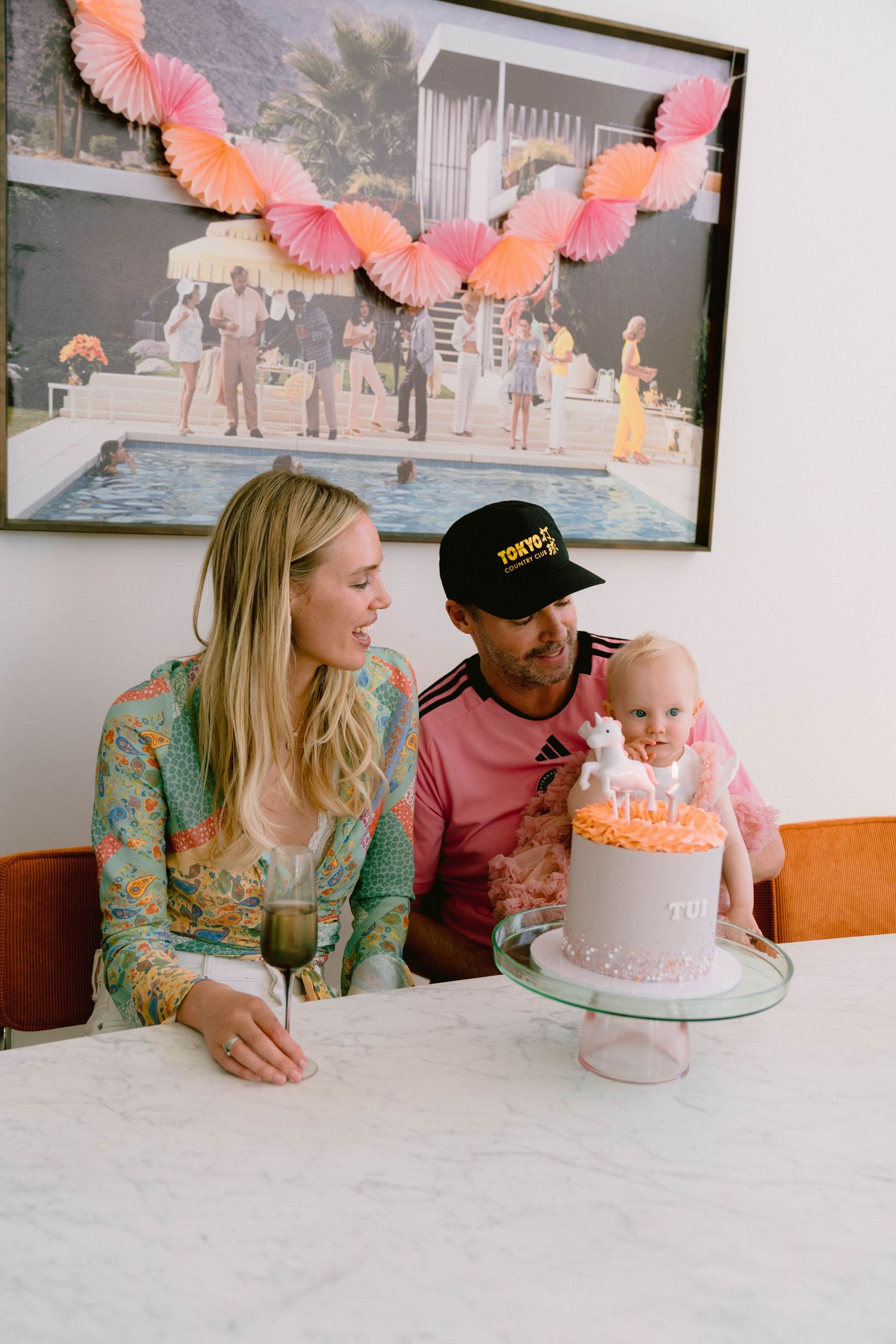 A celebratory moment around a pink and white birthday cake with coral paper decorations hanging above on the wall.