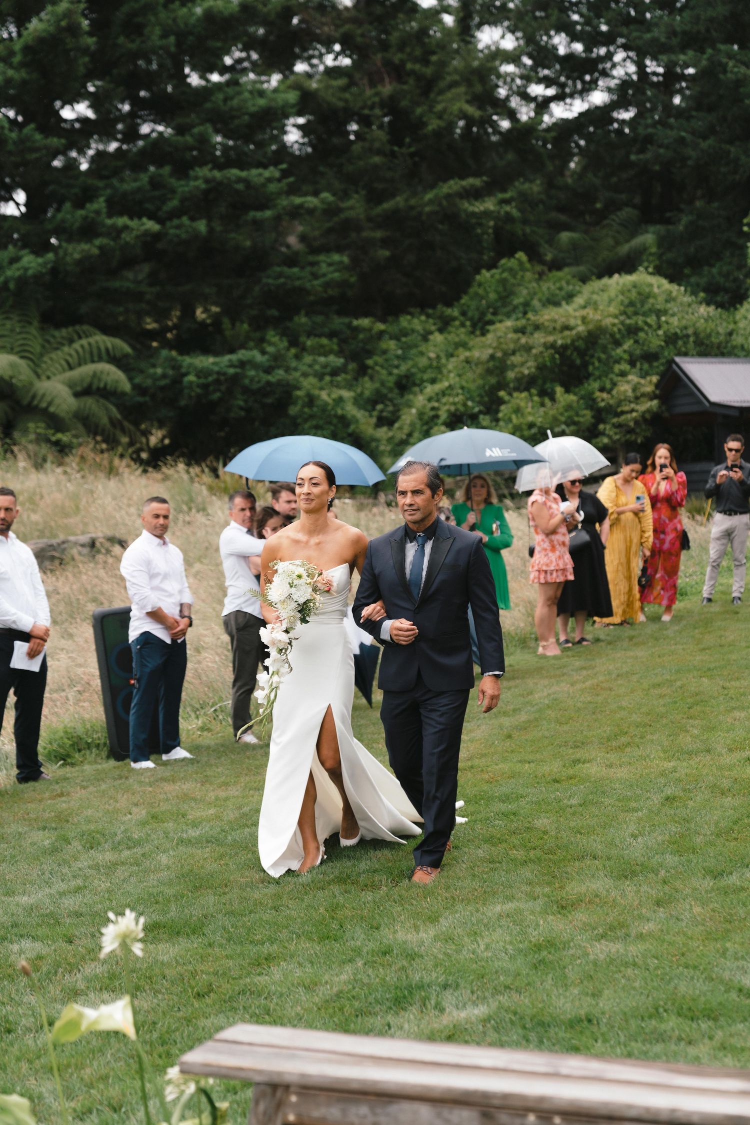Wedding party walking on a grassy hillside holding umbrellas during a light rain shower.