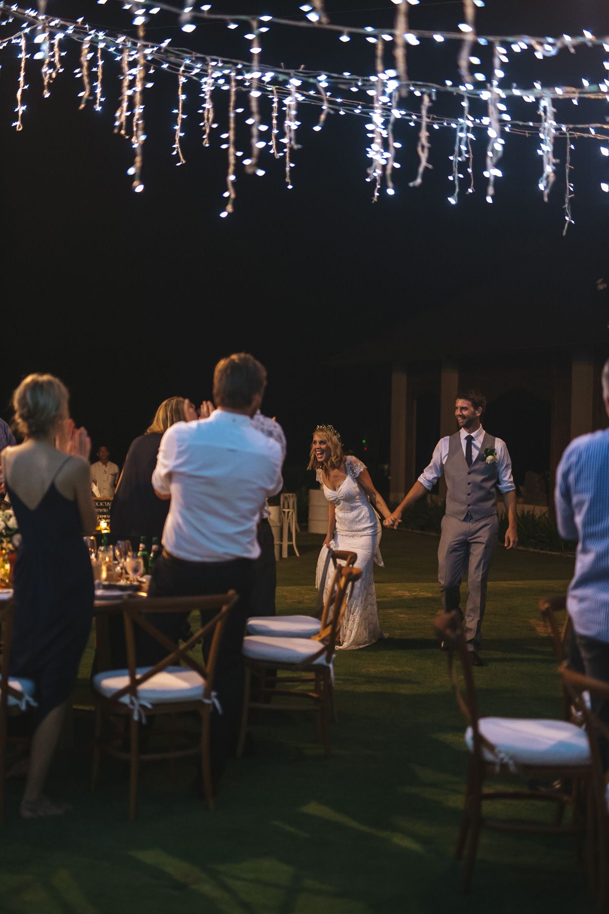 String lights illuminate an outdoor evening wedding reception as figures walk through rows of chairs on a lawn.
