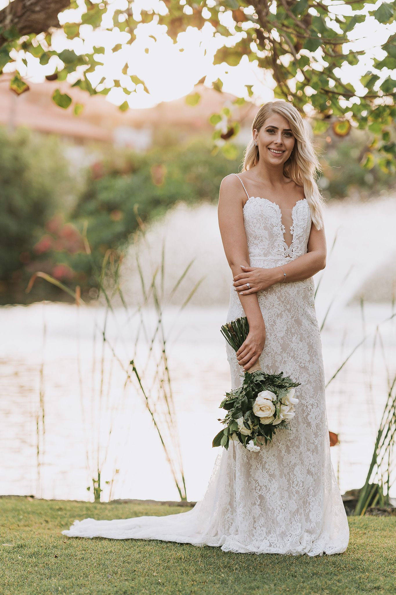 A white lace wedding dress is showcased in an outdoor bridal photo session near a lake during golden hour.