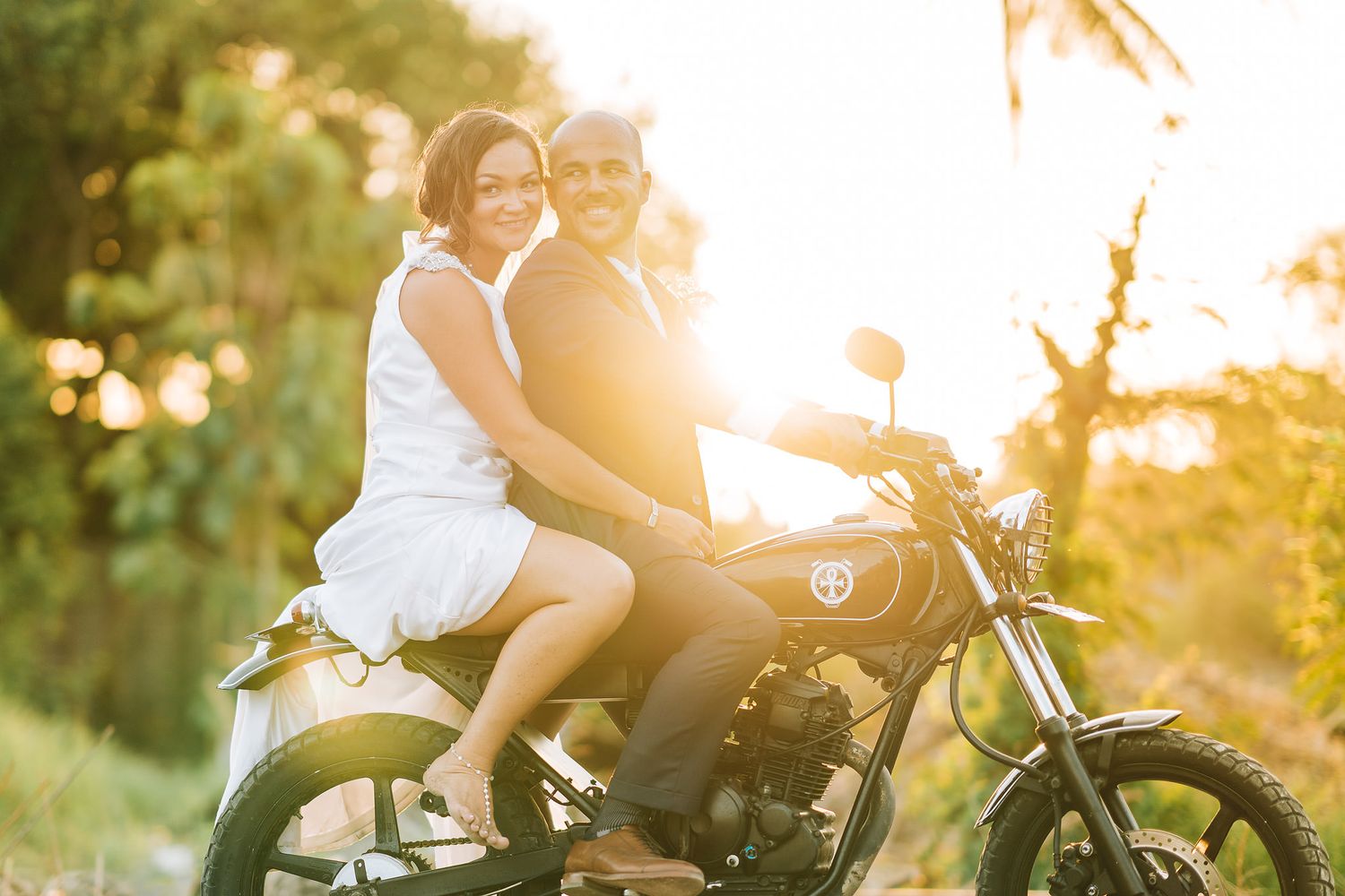 A couple sits on a motorcycle together against a warm sunset backdrop with golden bokeh lighting.