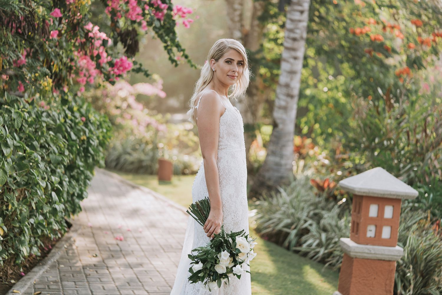 A bride in a lace gown holding a white bouquet poses in a lush tropical garden setting.