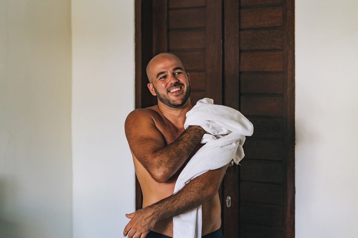 Shirtless person drying themselves with a white towel while standing near a wooden door in natural lighting.