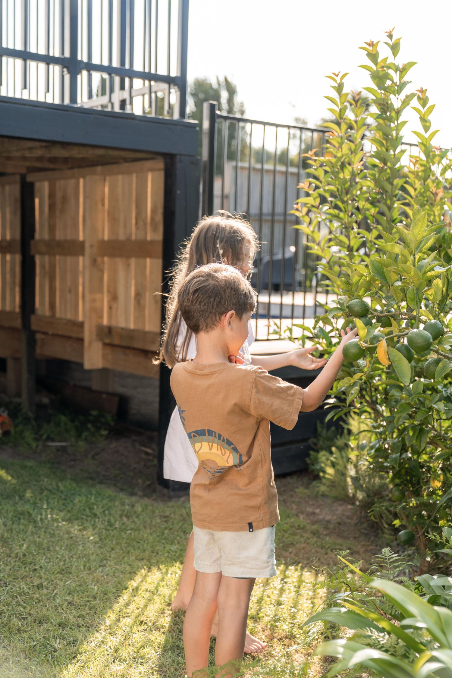 A toddler in a brown jacket explores the backyard garden while walking near green bushes and a wooden fence.