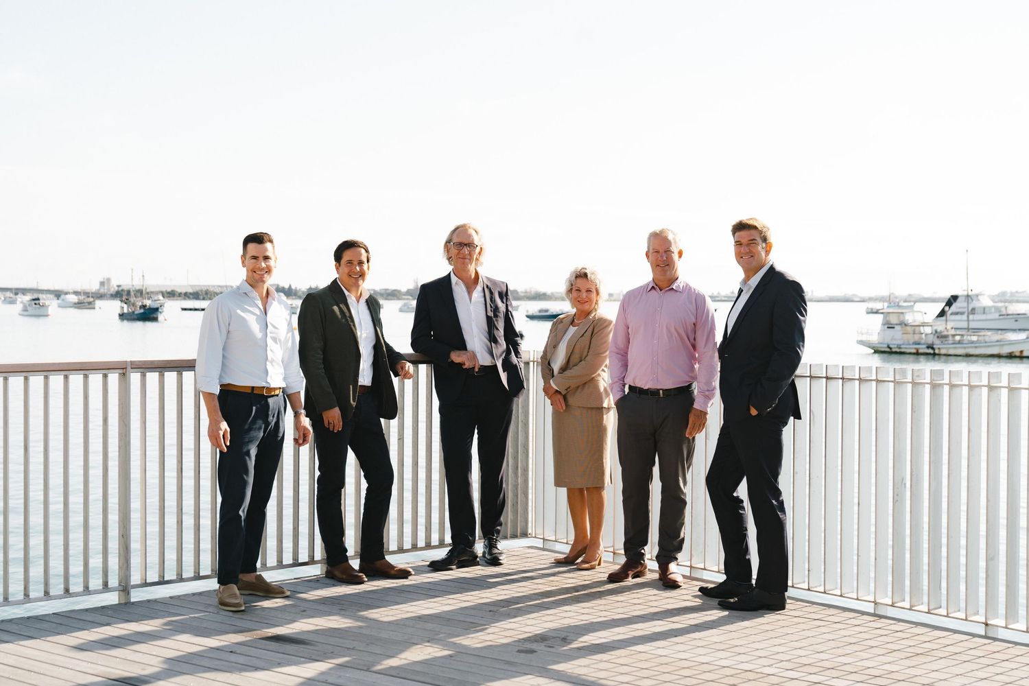 A group of formally dressed people standing together on a waterfront deck overlooking boats and water.