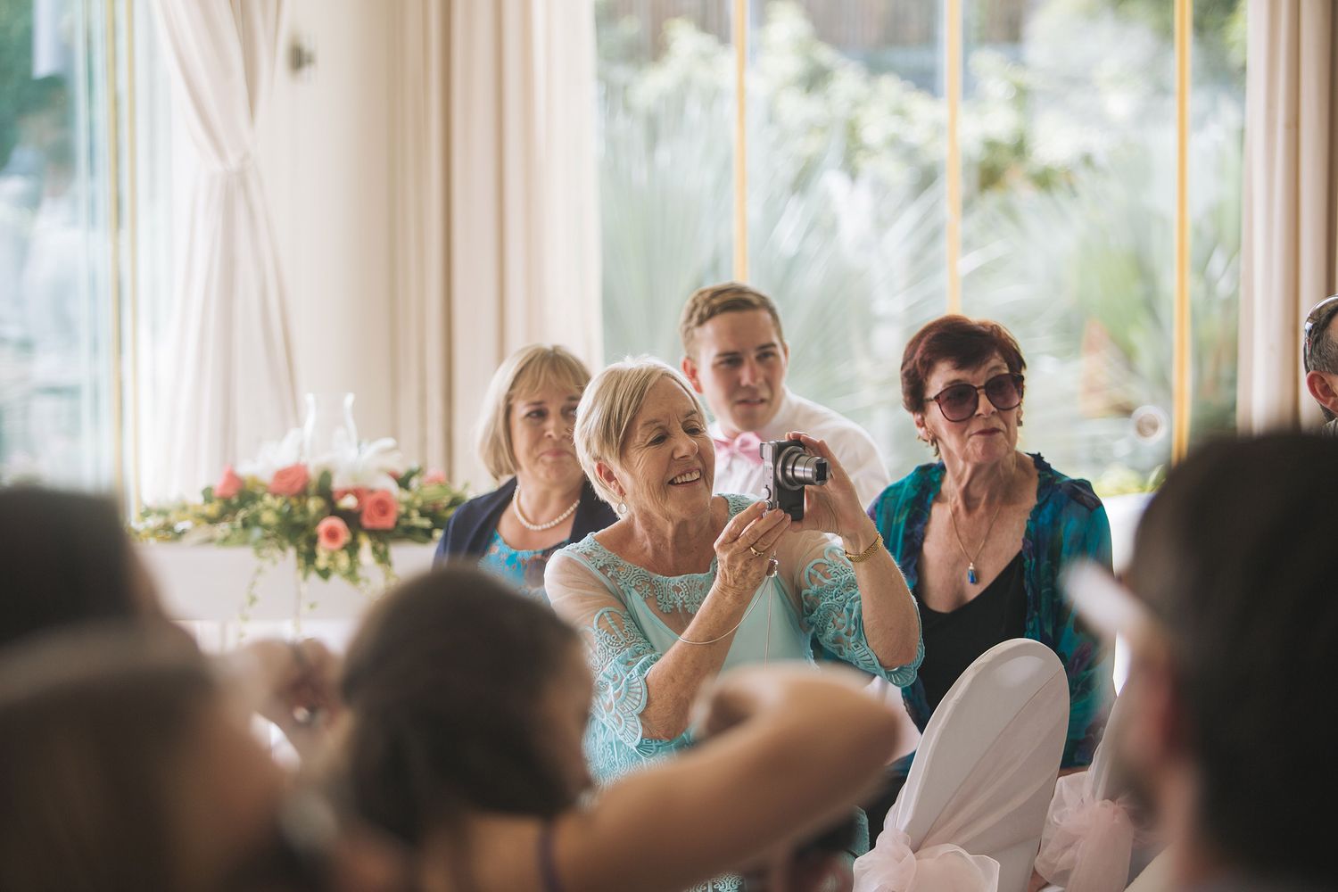 Wedding guests watching ceremony in sunlit room with large windows.