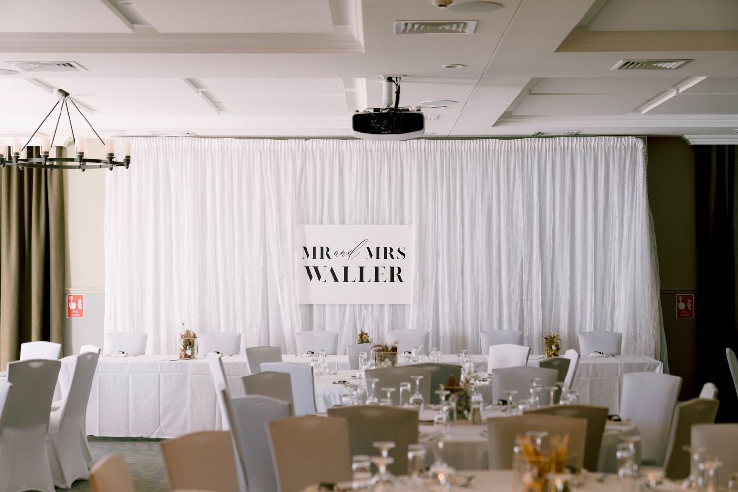 White wedding reception venue setup with elegant draped backdrop displaying 'Mr & Mrs Waller' signage and round tables.