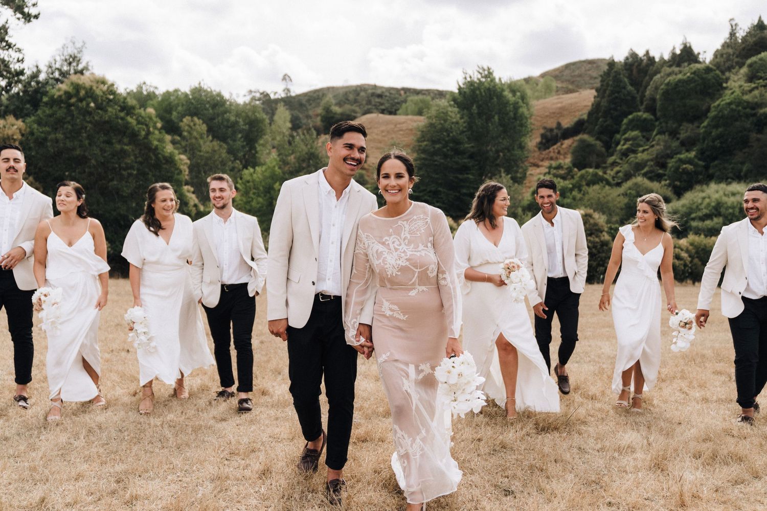 Wedding party walking together on a grassy hillside wearing coordinated white and cream outfits.