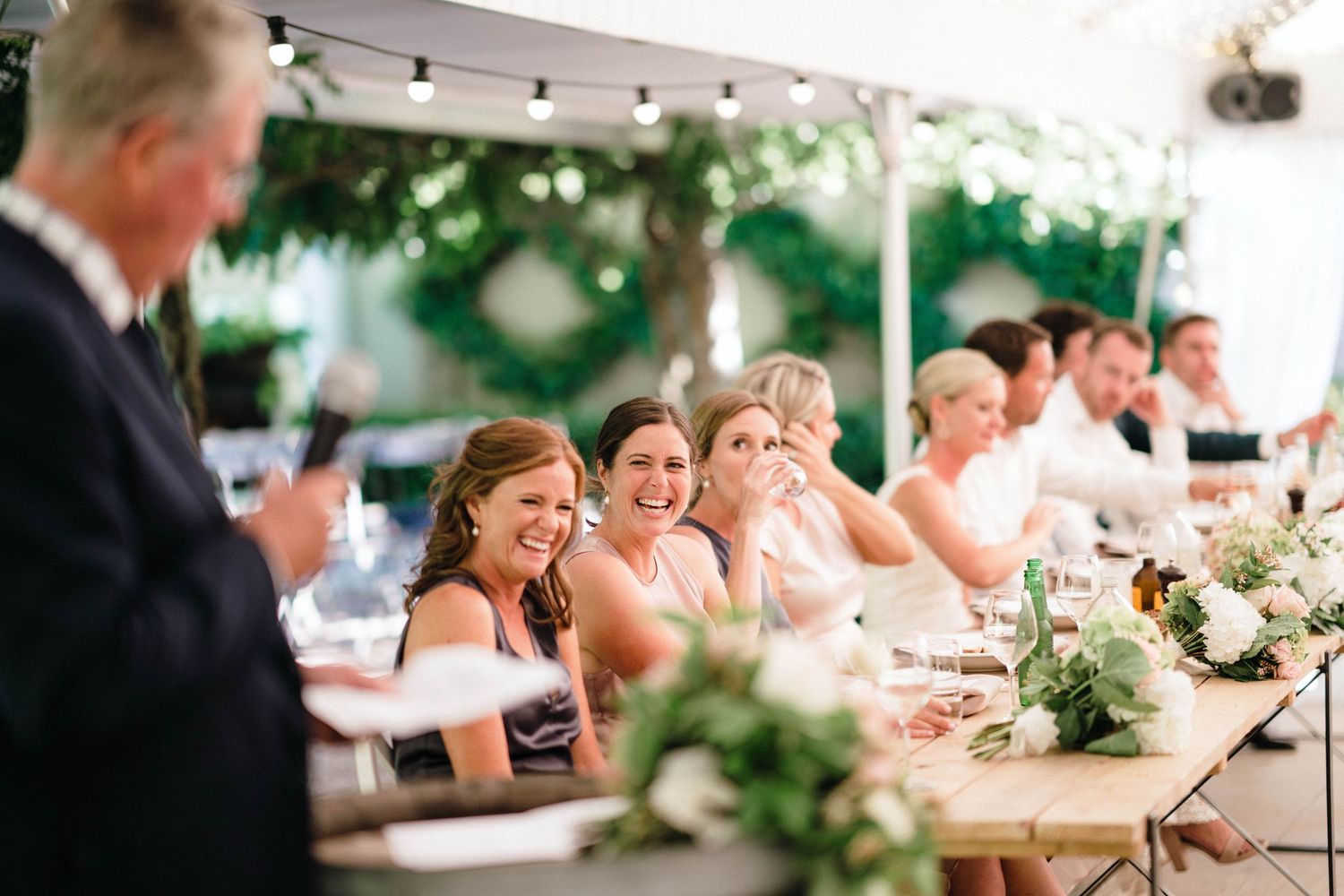 Wedding guests enjoy an outdoor reception dinner under string lights at long tables with greenery decor.