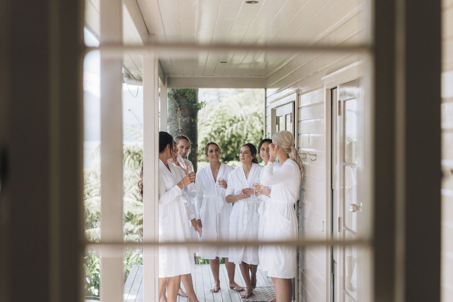 A group in white dresses gathers on a sunlit porch during wedding preparations.