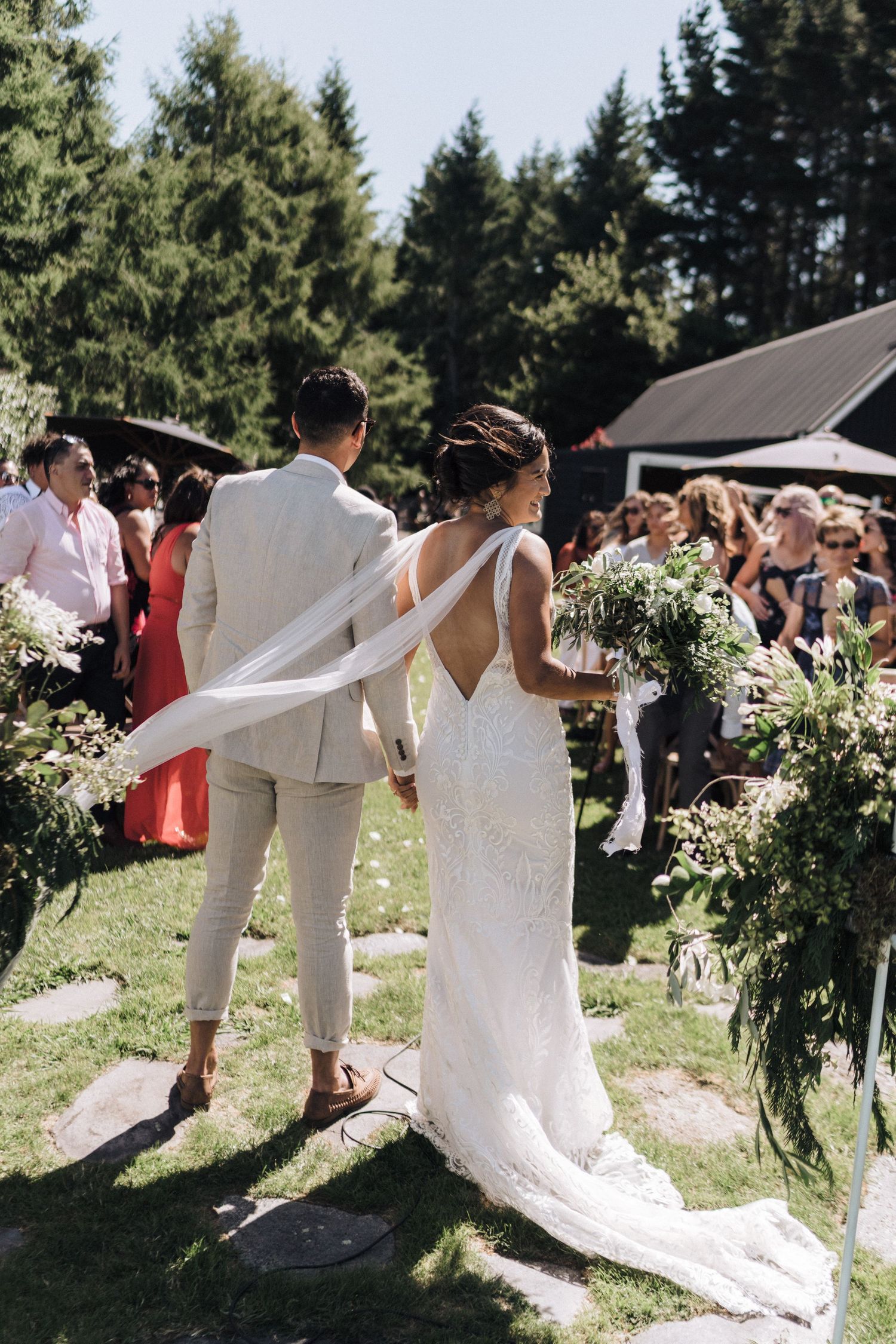 A wedding couple stands before guests in a sunlit garden ceremony with lush evergreen trees in the background.