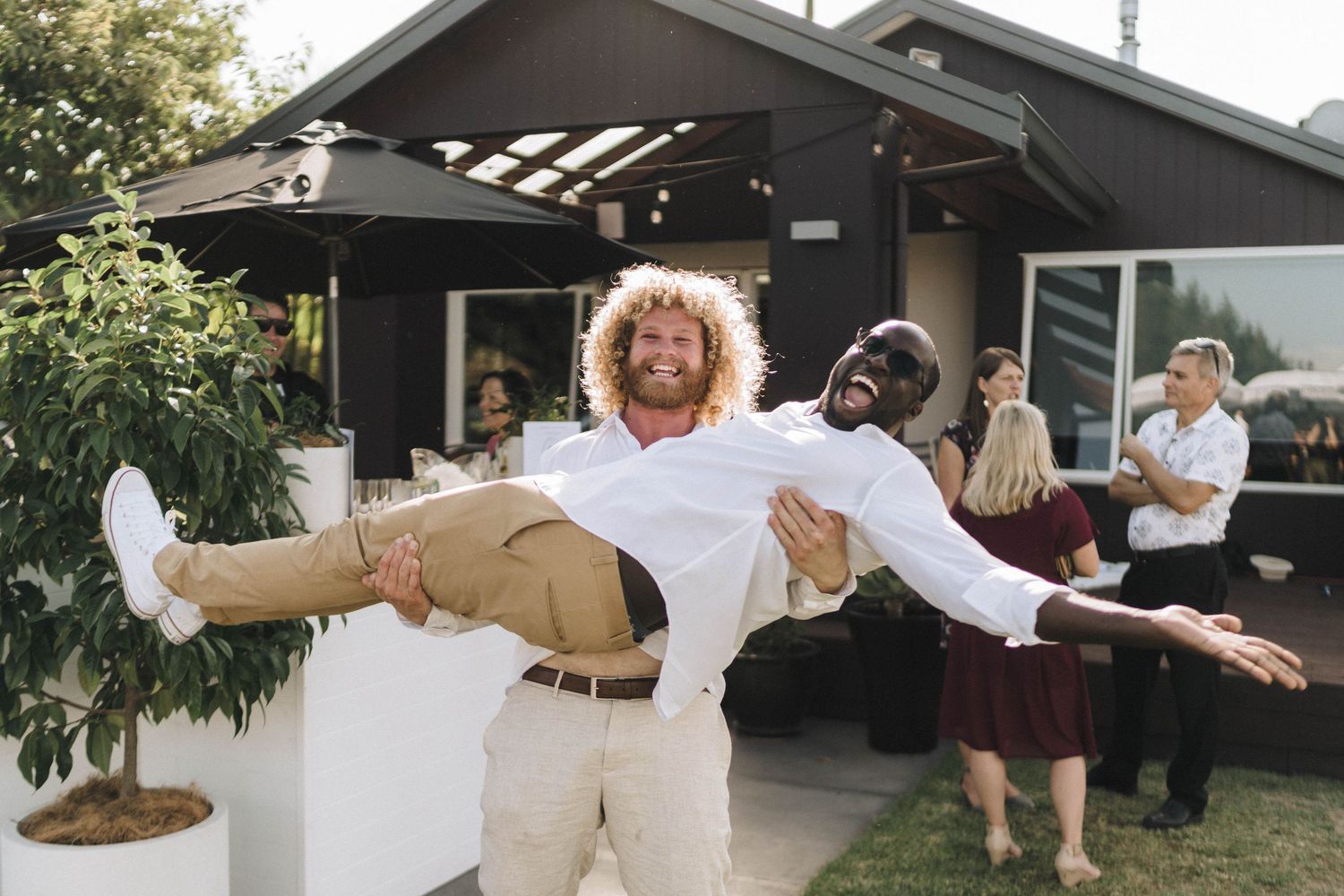 Playful wedding guest performs lift pose while others watch at outdoor celebration.