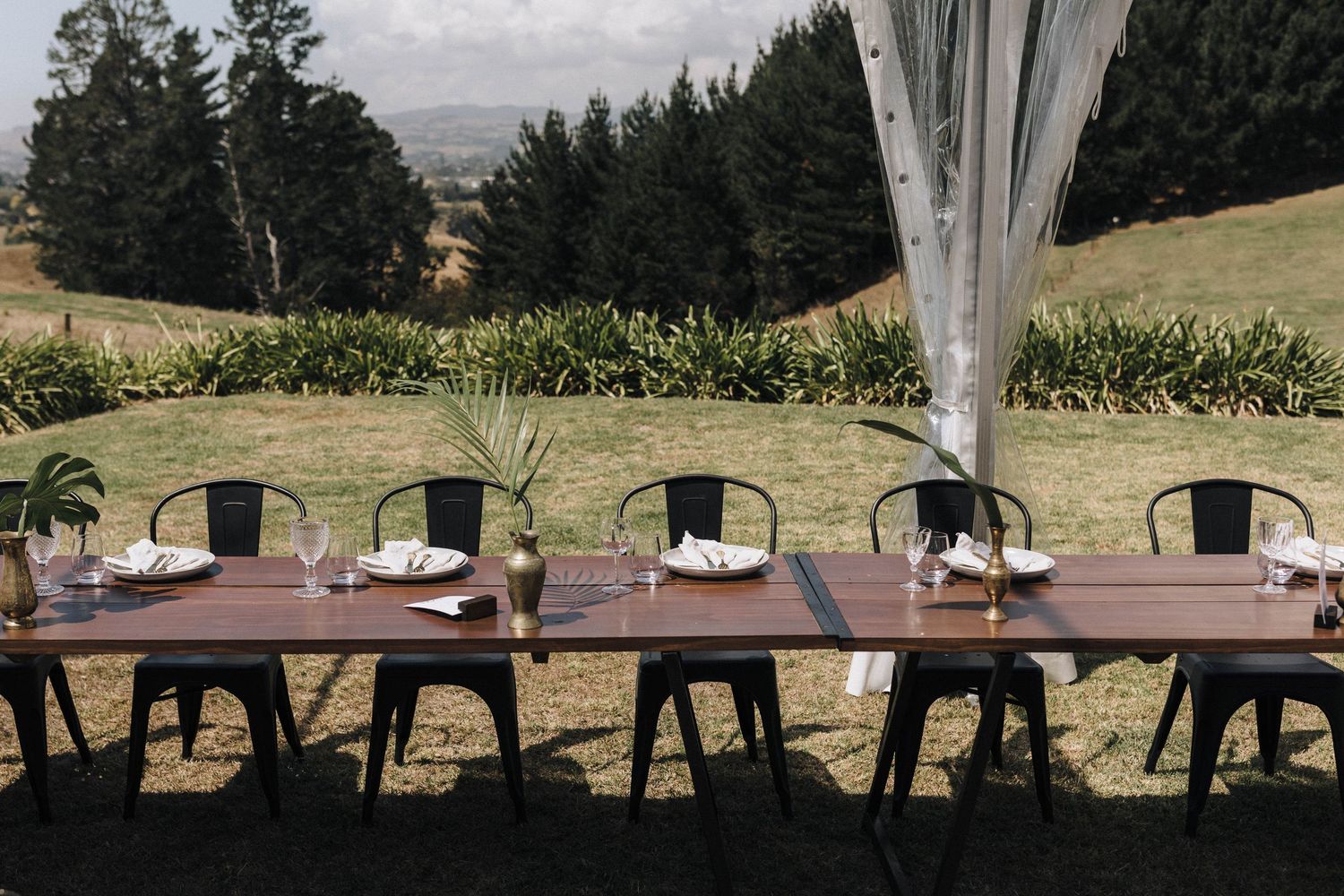 Long rustic wooden tables set for an outdoor wedding reception under strung lights and tall evergreen trees.