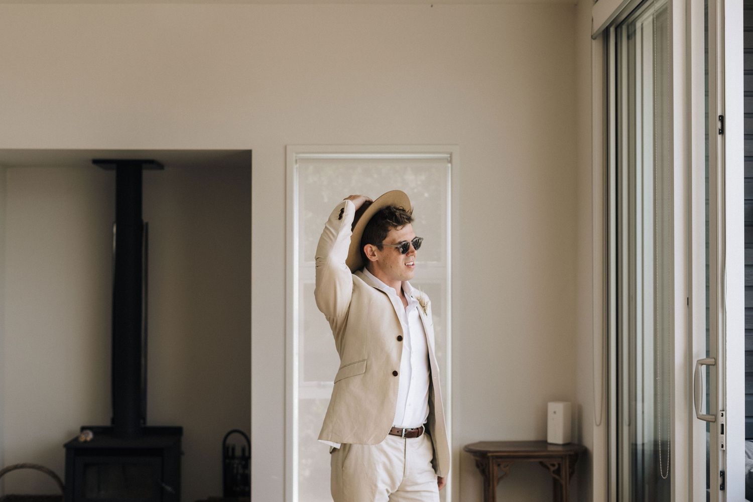 Stylish person in a cream colored suit and sunglasses poses against a light wall in a minimal interior space.