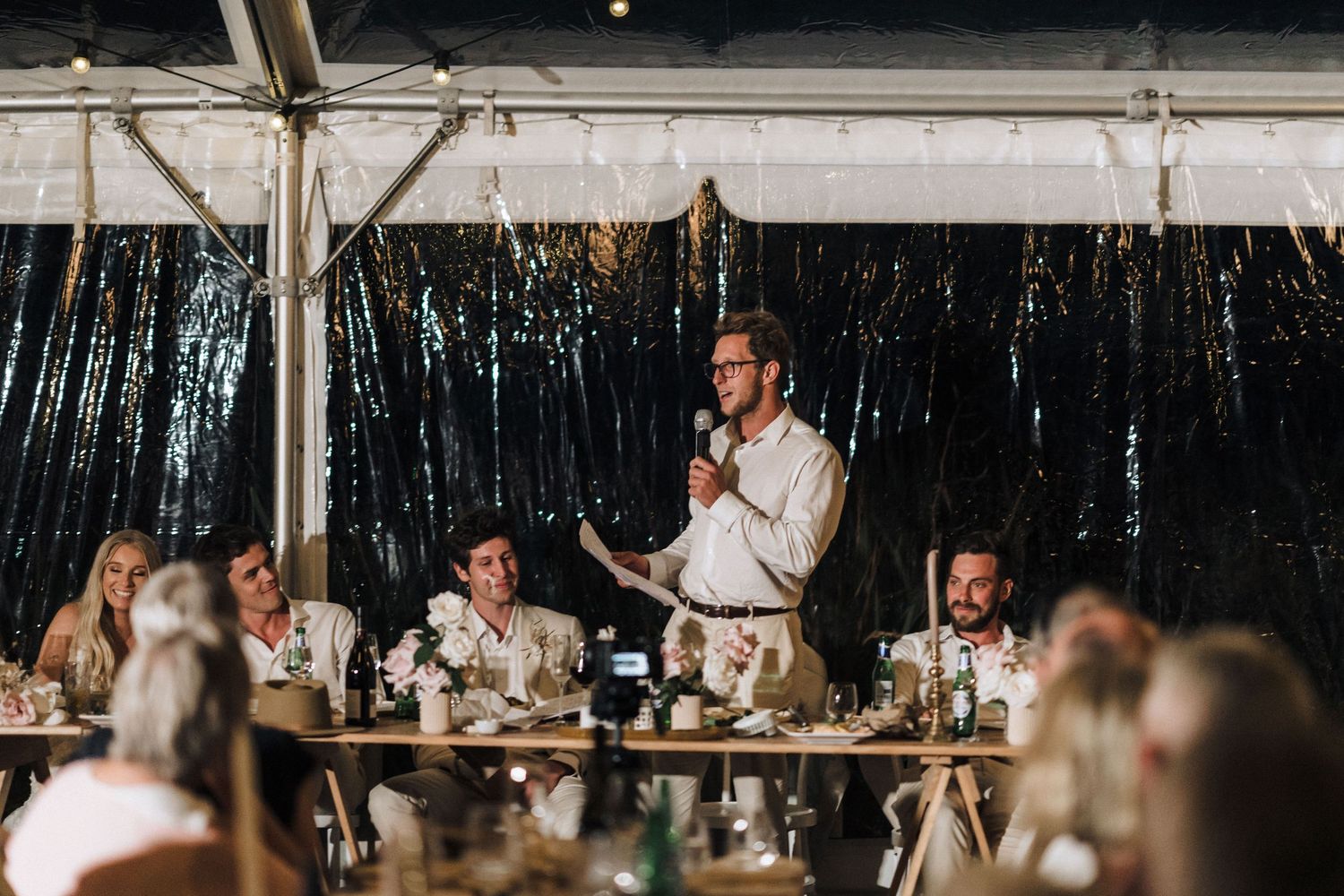 Wedding guests sit at a long reception table in a rustic venue with dark curtains and ambient lighting.