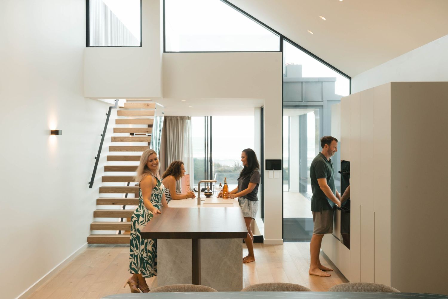 People gather around a contemporary kitchen island in a modern home with wooden stairs and high ceilings.