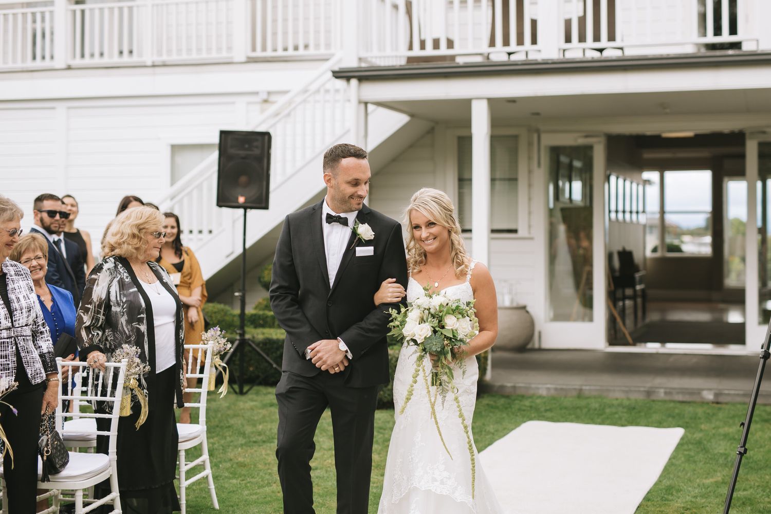 Two people celebrate their wedding ceremony outdoors in front of a white colonial style building while guests look on.