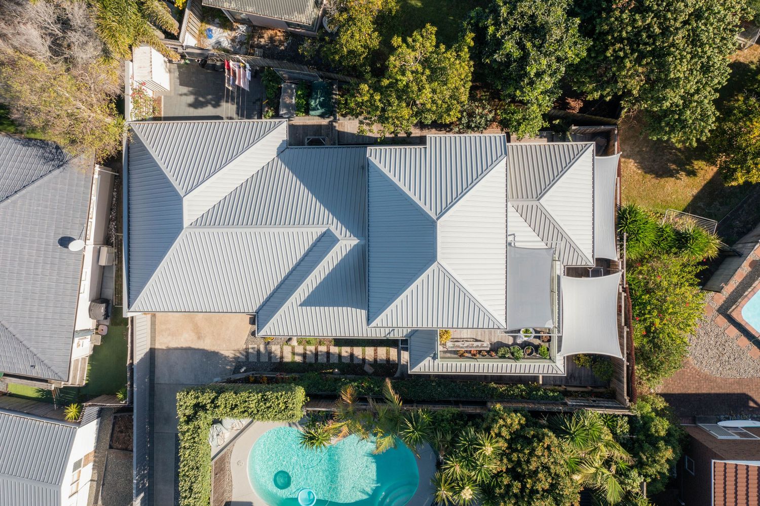 Aerial drone view of modern house with white metal roof and turquoise swimming pool surrounded by green trees.