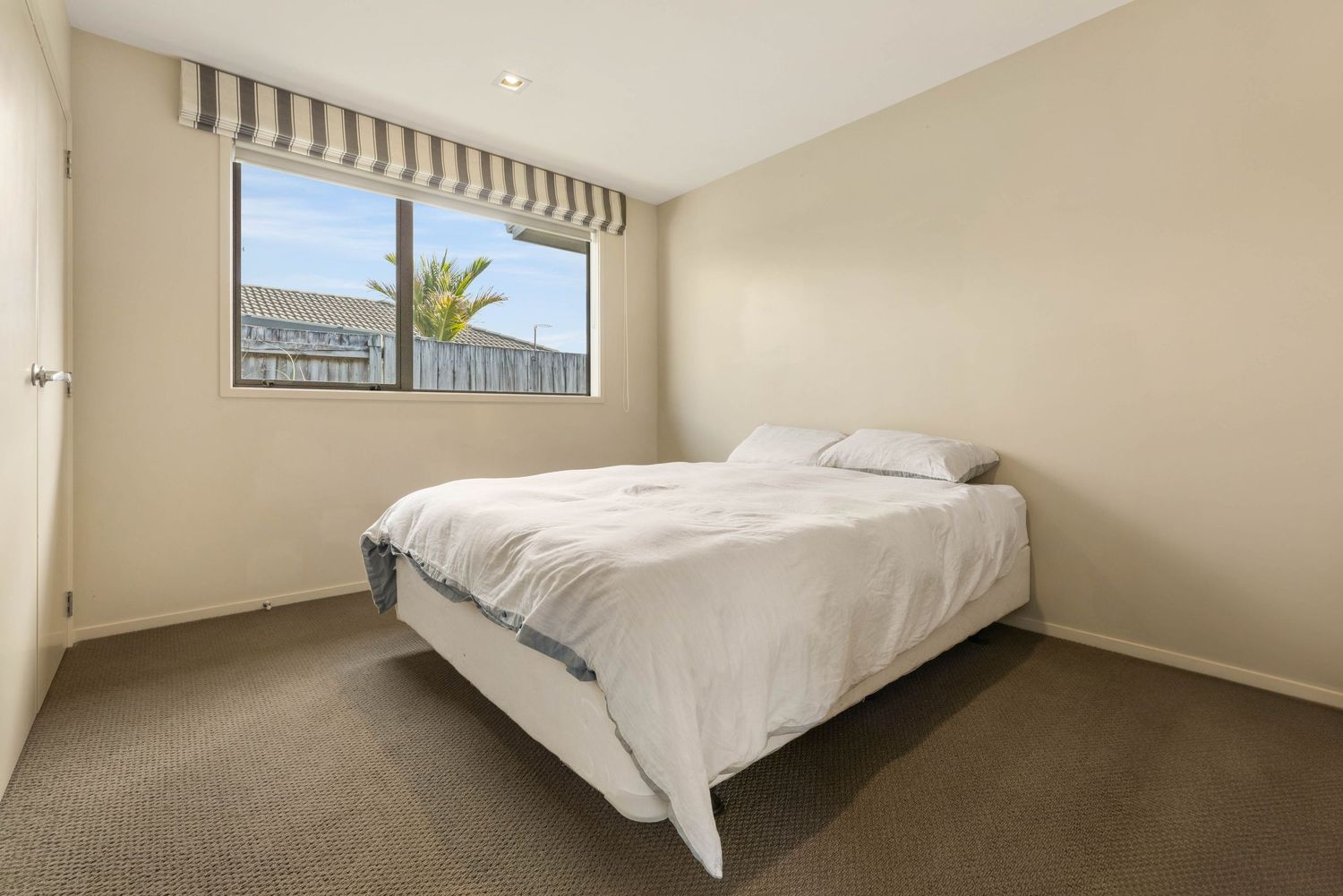 Minimalist bedroom with white bedding, striped roller blind, and carpet flooring bathed in natural light.