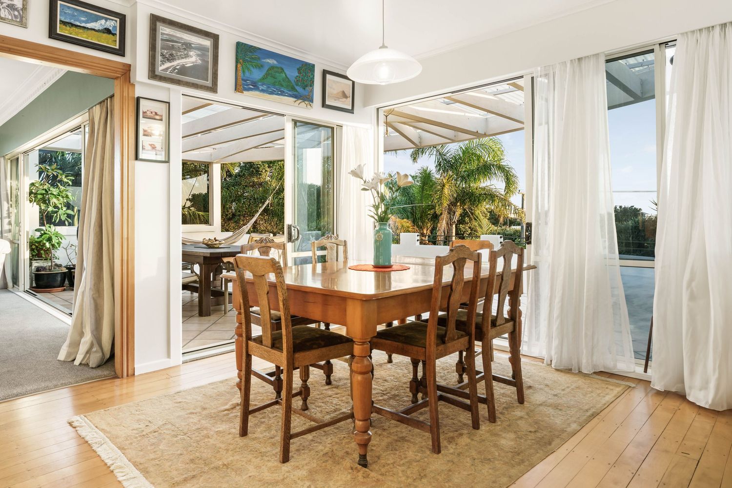 Bright coastal dining room with wooden table and chairs surrounded by floor-to-ceiling windows and white curtains.