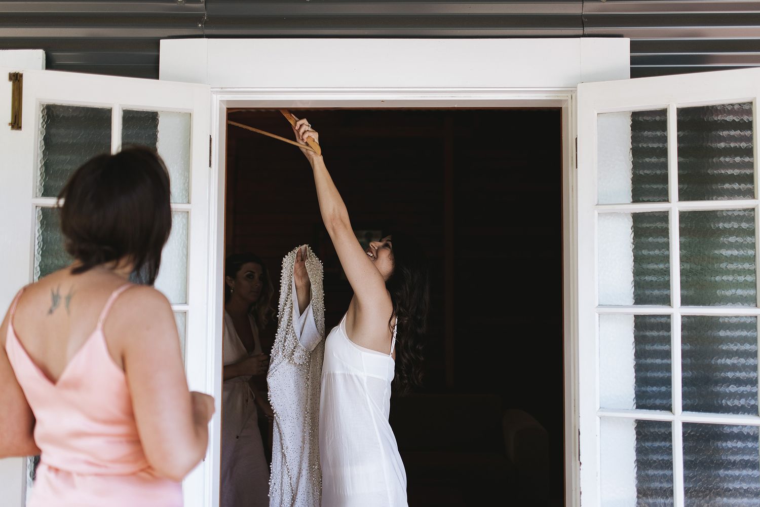 Two figures stand in a doorway with white trim, one wearing pink and another with raised arm against dark interior.