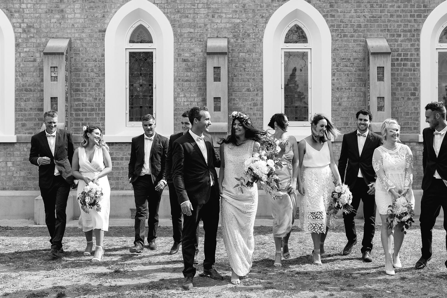 Wedding party walking together outside brick church with arched windows in black and white photography.