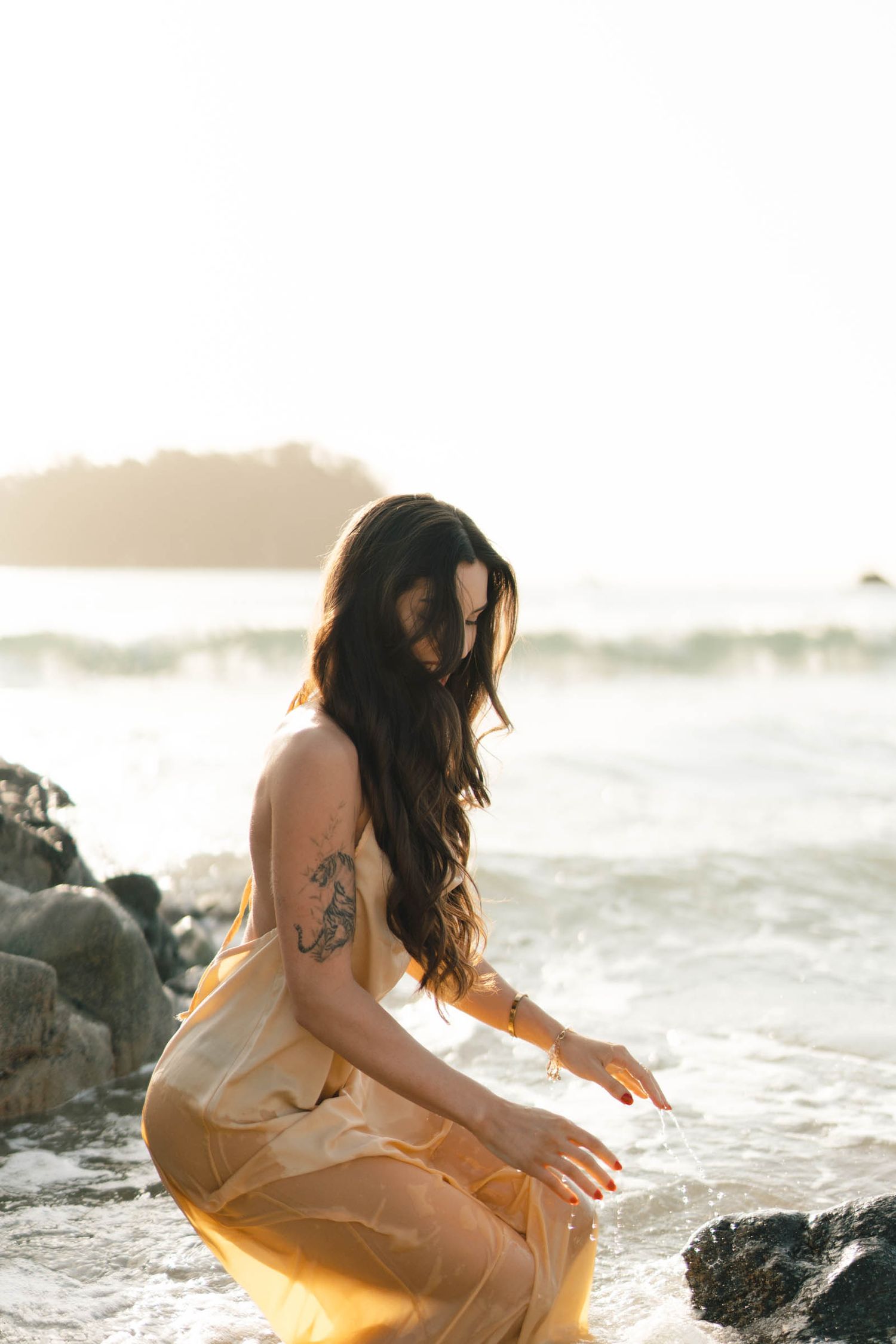 Soft beige dress contrasts with dark rocks as waves crash during golden hour.