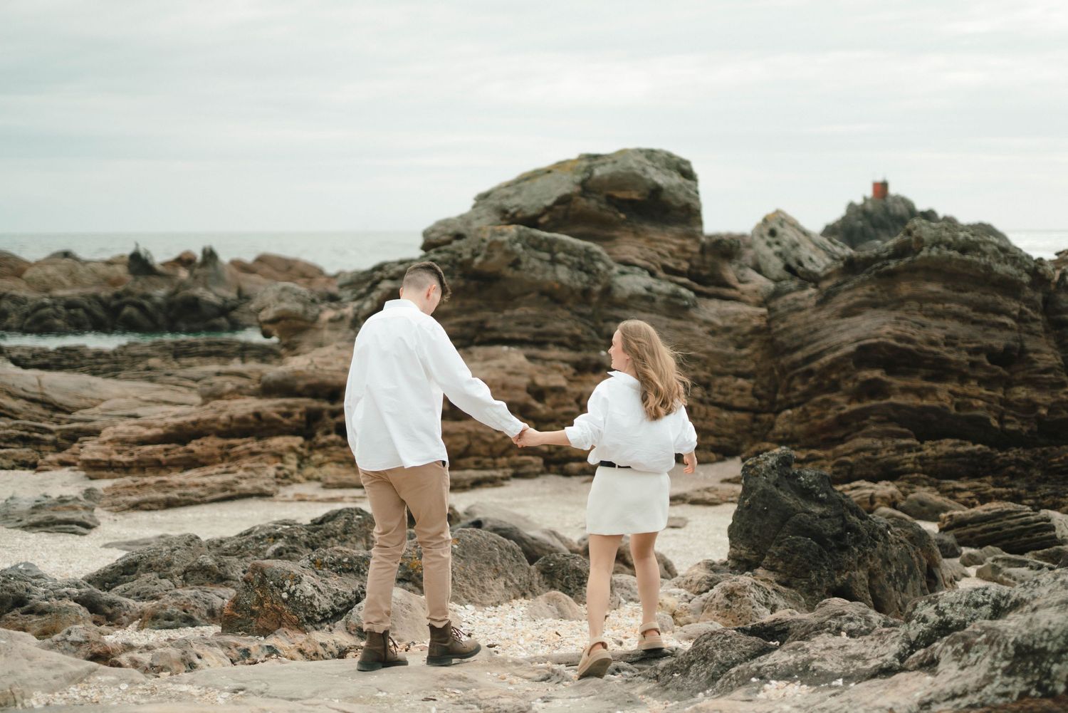 Two people in light-colored outfits walking hand-in-hand along dramatic rocky coastal cliffs with ocean views at sunset.