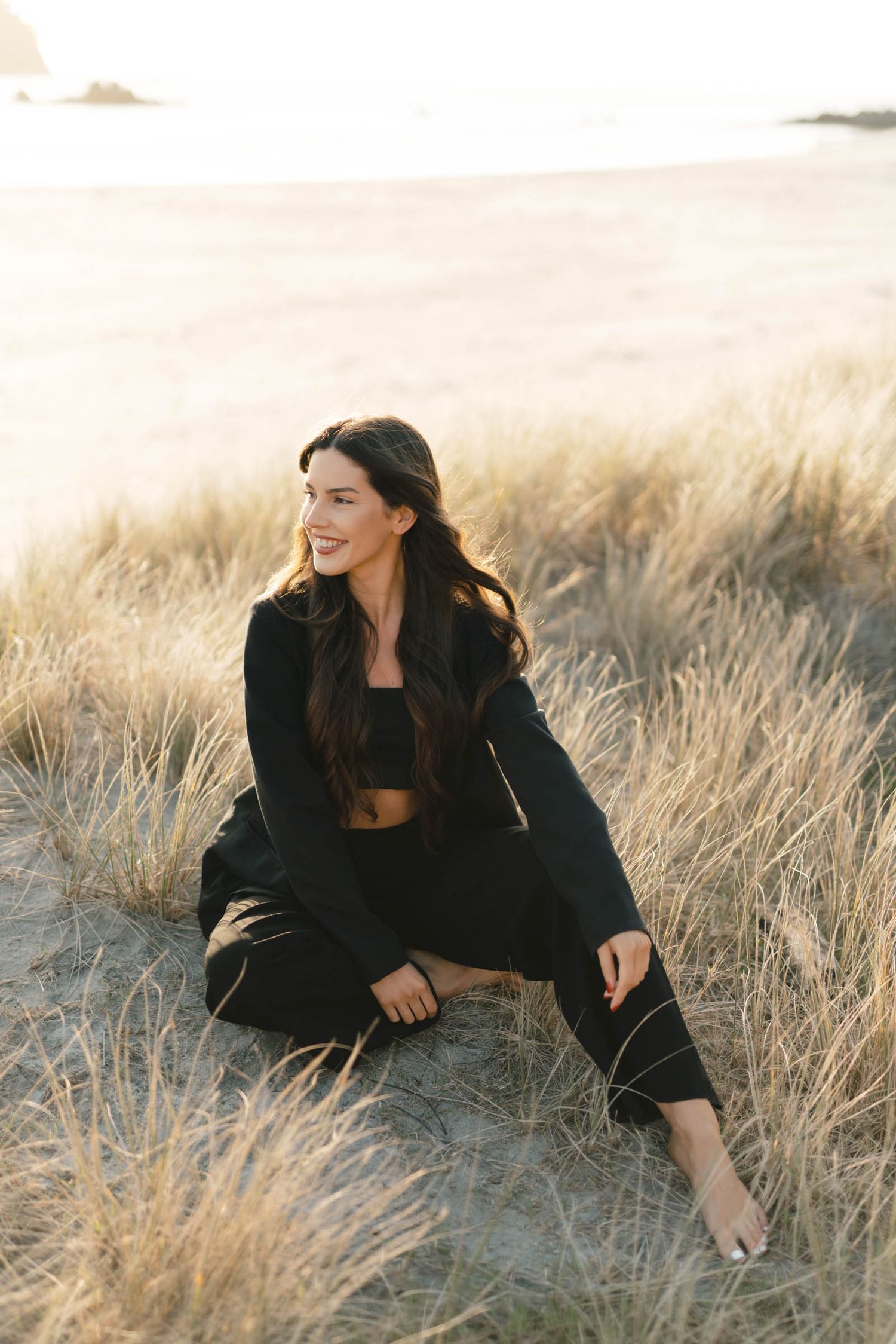 Model in black outfit poses playfully in golden beach grass during sunset photoshoot.