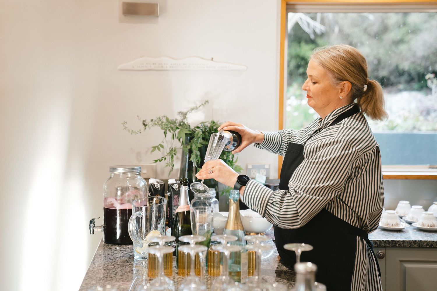 Bartender pouring drinks at a counter with glassware and botanicals in the background.