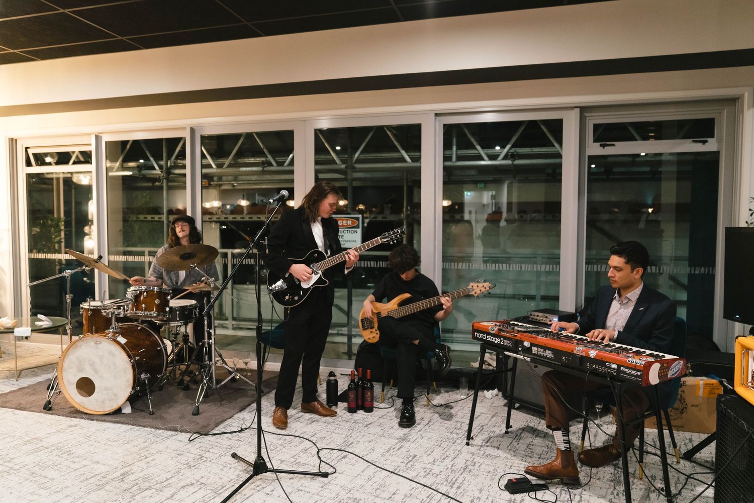 Live band performing with instruments including drums and guitar in front of glass windows.